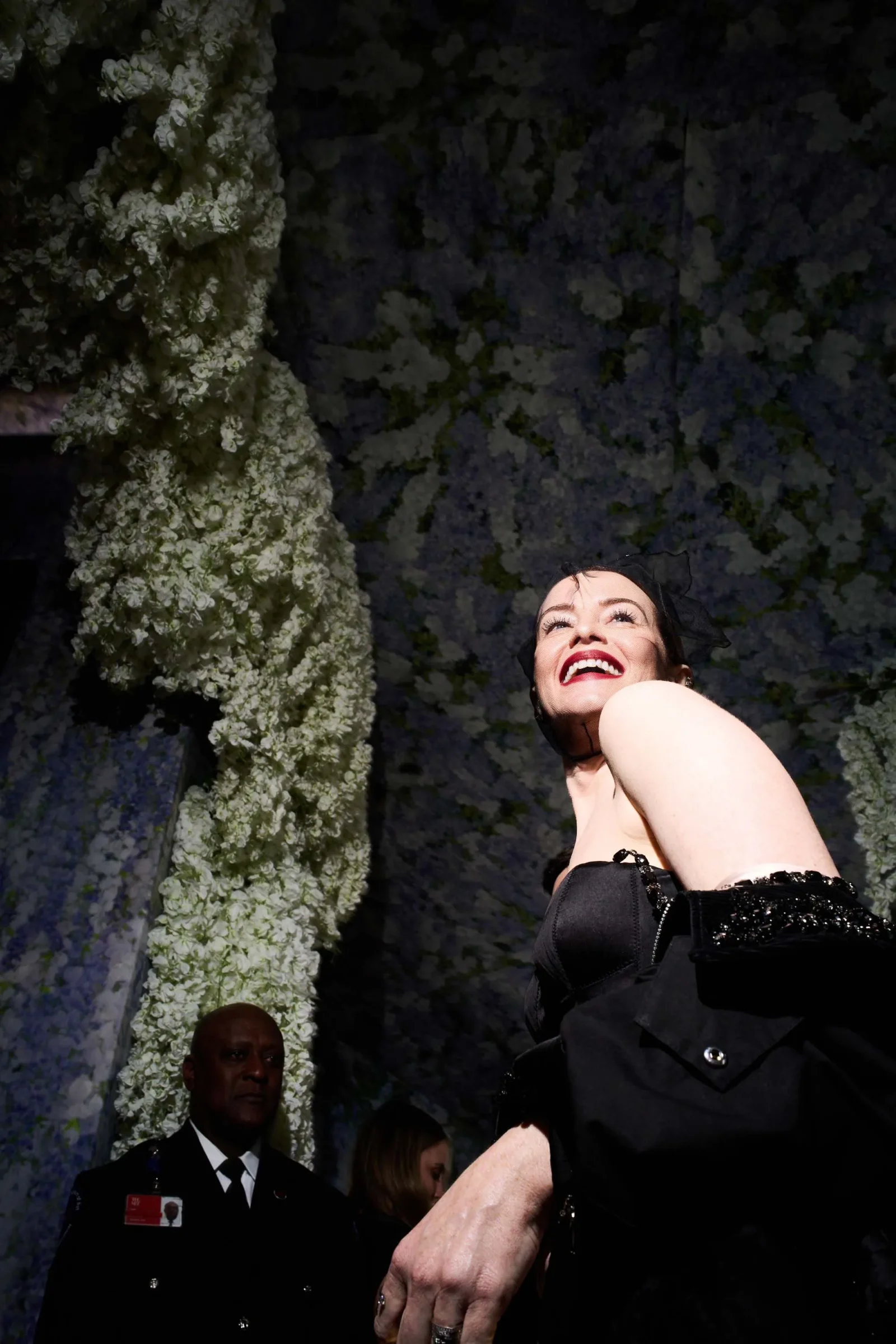 Smiling woman in a black evening gown poses against a wall of cascading white flowers, with a security guard in a dark uniform visible in the background.