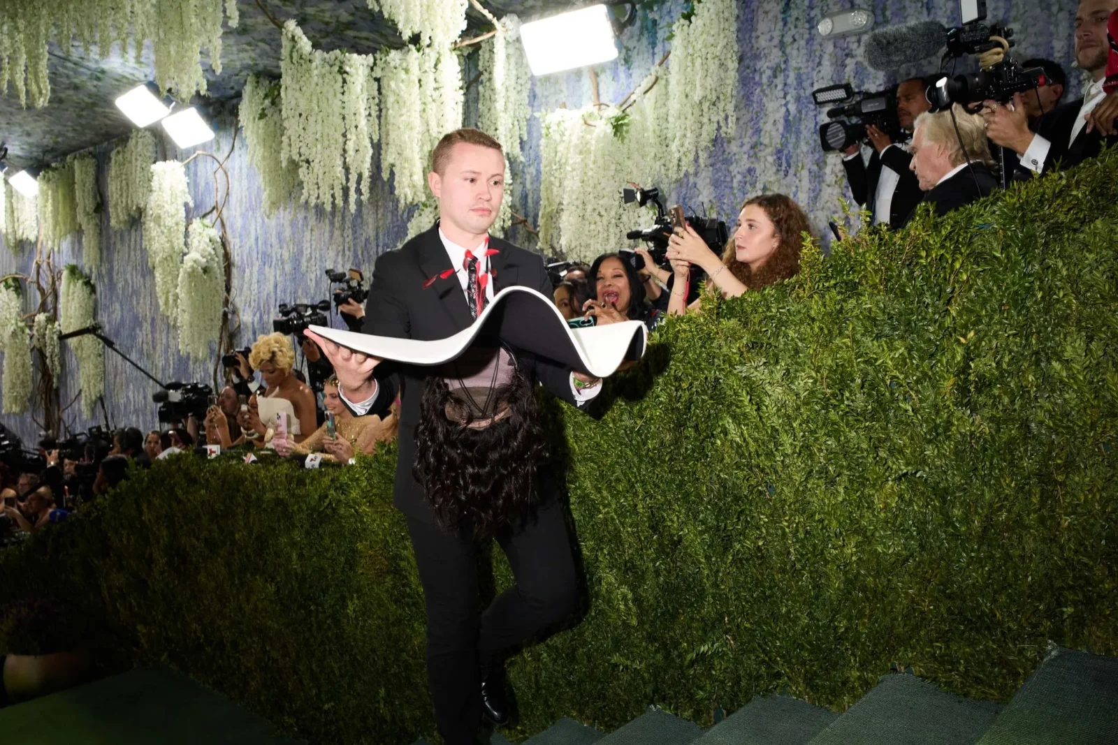 Man in a black suit and red bow tie holding a large open book or award on a lush, floral backdrop while photographers capture the moment.