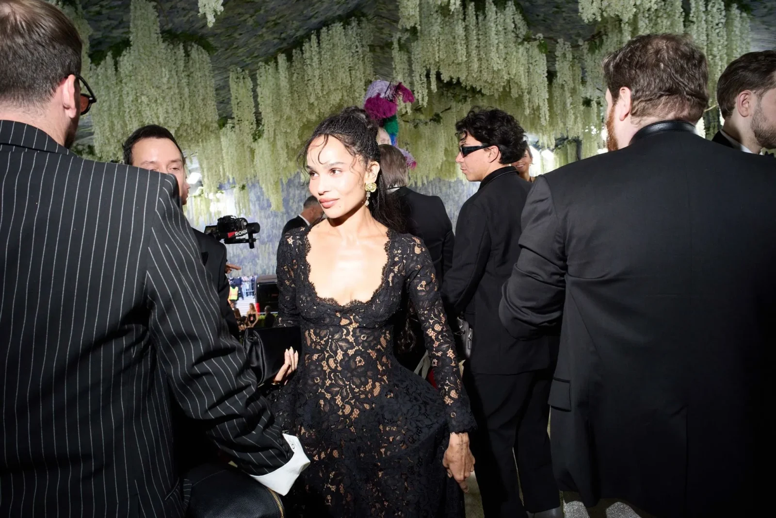 Woman in a black lace dress stands among formally dressed guests at an event, with hanging white flowers overhead.
