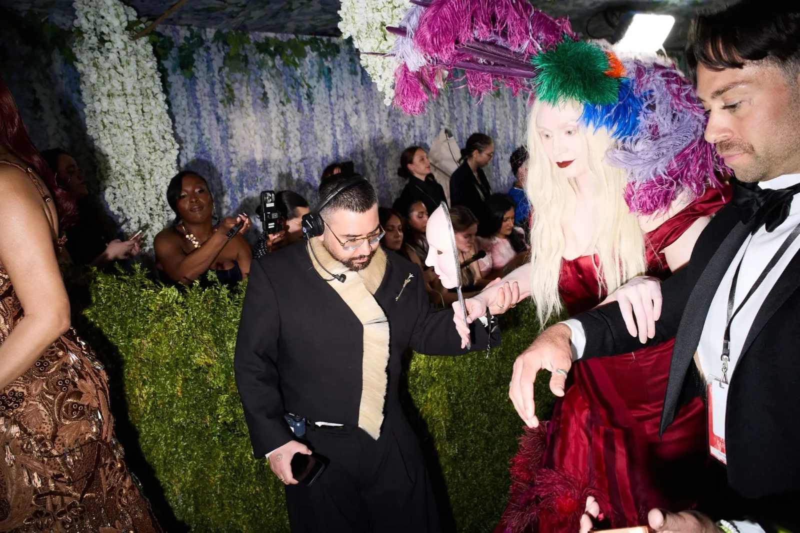 Backstage at a fashion show with guests and models mingling; a blonde woman wears a bright multicolored feathered headpiece against a floral backdrop while others in formalwear surround her.