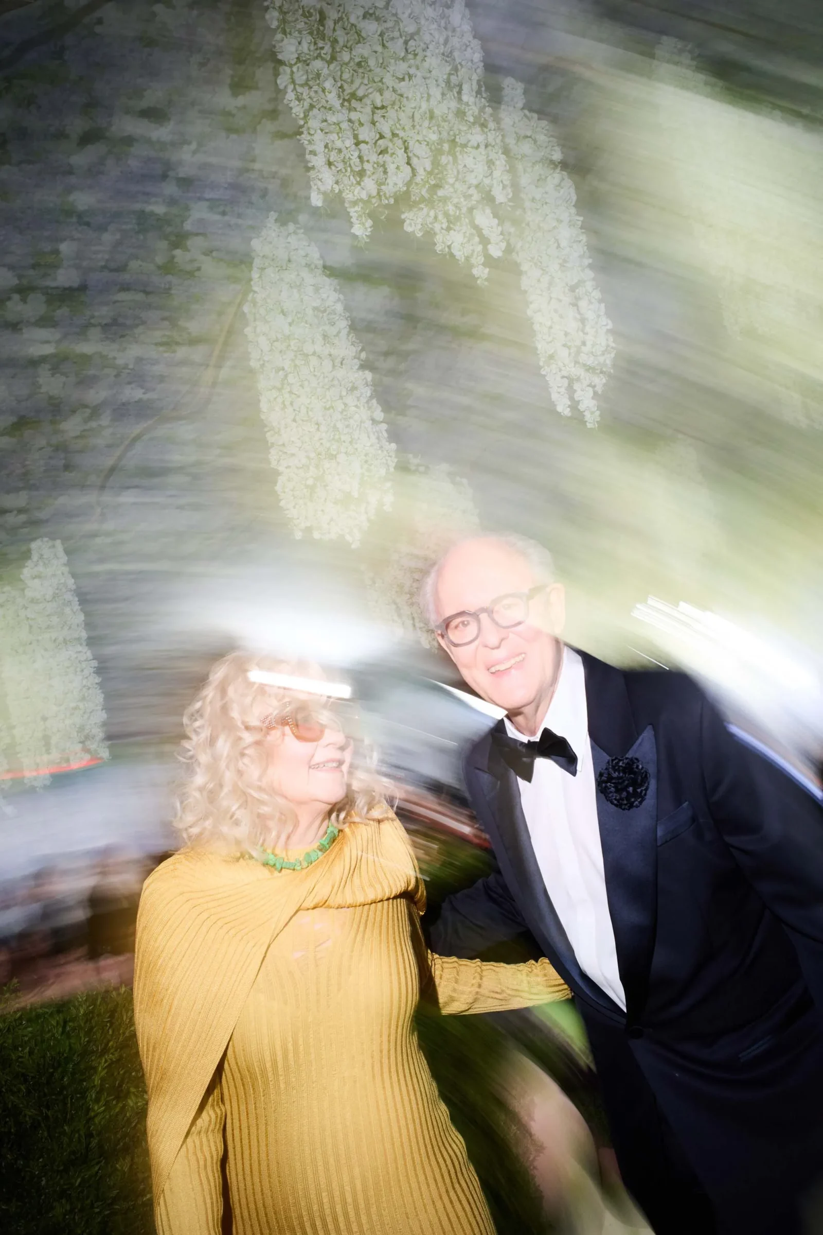 Smiling couple dancing at a formal event; woman in a mustard yellow ribbed dress with a green necklace, man in a tux, motion blur around them.