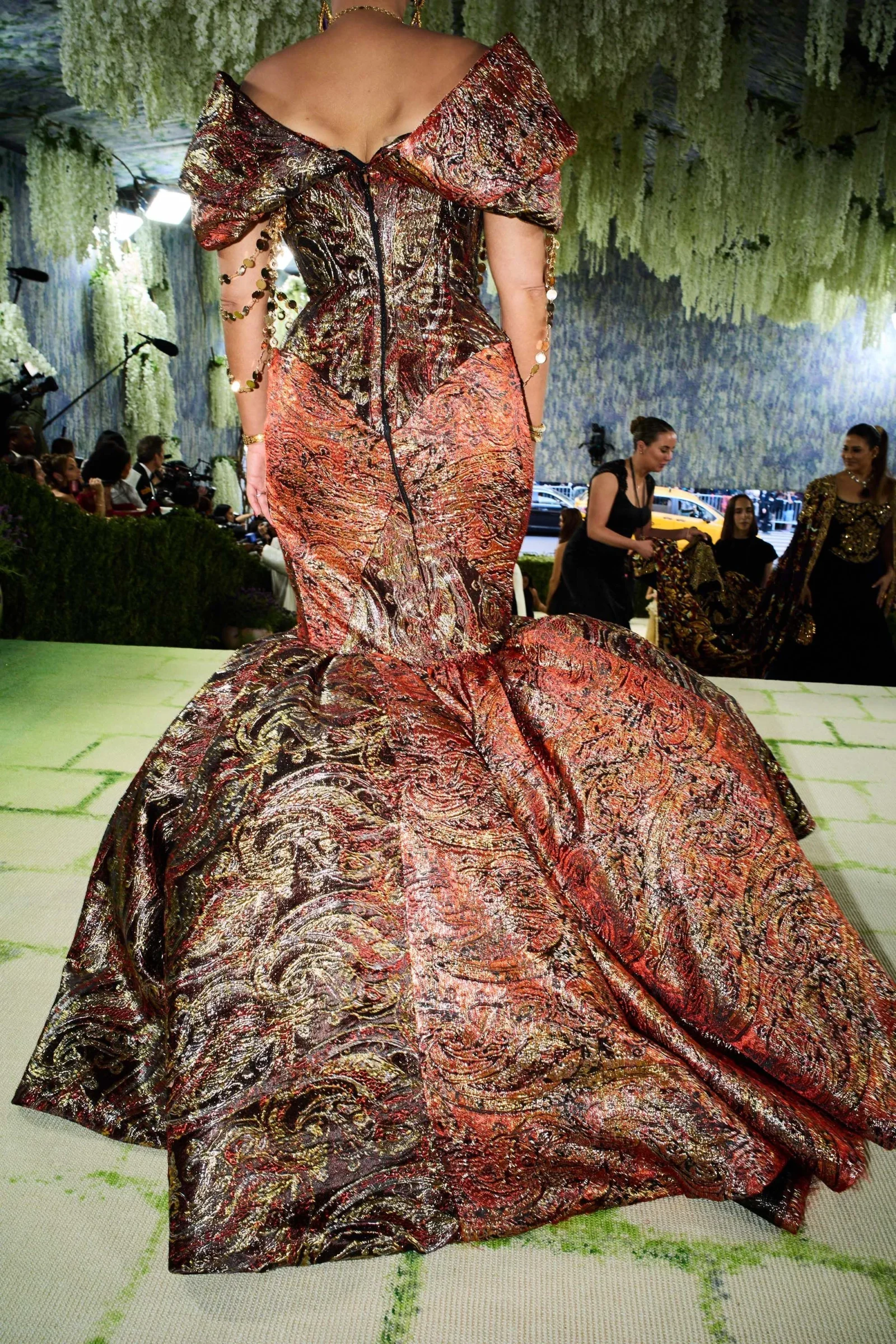 Back view of a model wearing a shimmering red-gold brocade ball gown with off-the-shoulder sleeves on a fashion runway