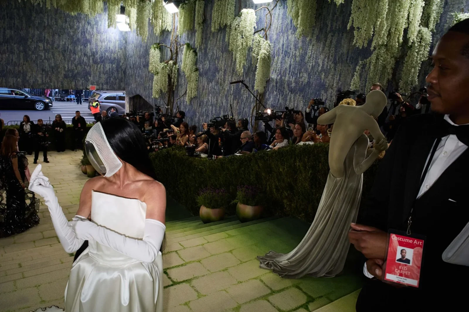 Celebrities posing on a green-carpeted red carpet with photographers and a hanging floral ceiling in the background.