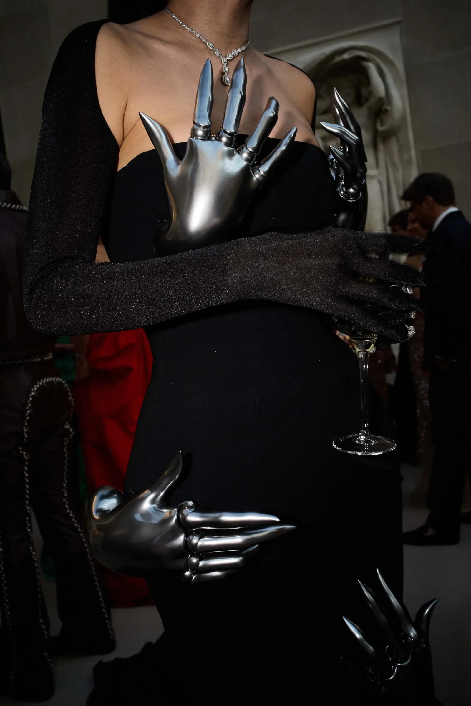 Woman in a black off-shoulder gown with silver claw-like prosthetic gloves, holding a champagne flute and wearing a diamond necklace.
