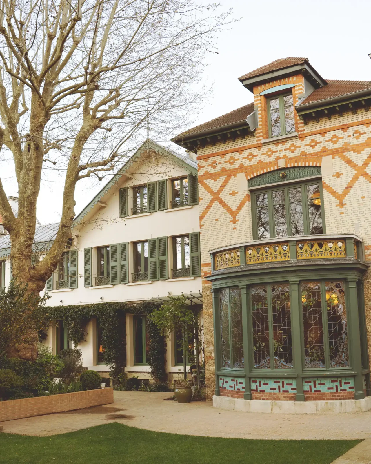 Brick house with decorative orange and white brick pattern, green window frames, and a rounded green conservatory-style bay window; bare trees in foreground.
