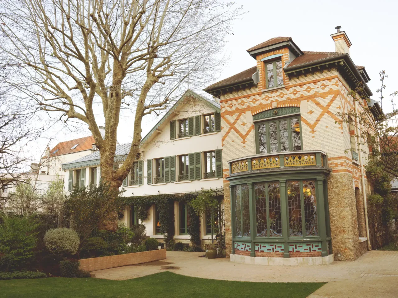 Brick house with decorative orange and white brick pattern, green window frames, and a rounded green conservatory-style bay window; bare trees in foreground.