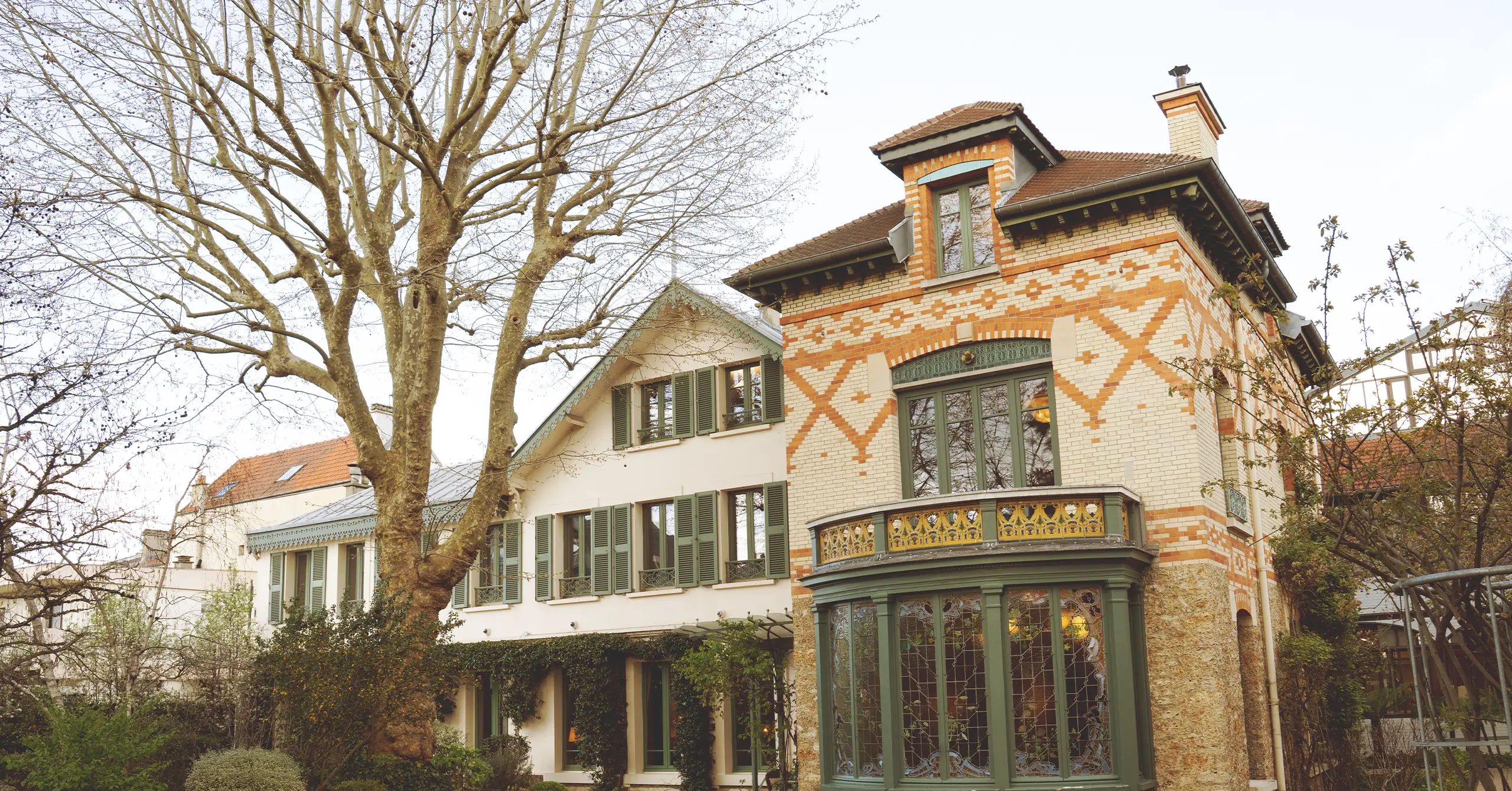 Brick house with decorative orange and white brick pattern, green window frames, and a rounded green conservatory-style bay window; bare trees in foreground.