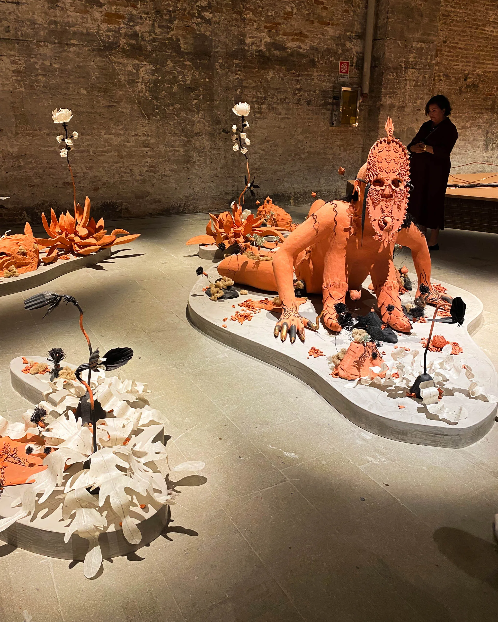 Peach-colored fantasy creature sculpture with an ornate mask on a circular pedestal, surrounded by orange plant-like forms in a brick-walled gallery; a viewer stands nearby.