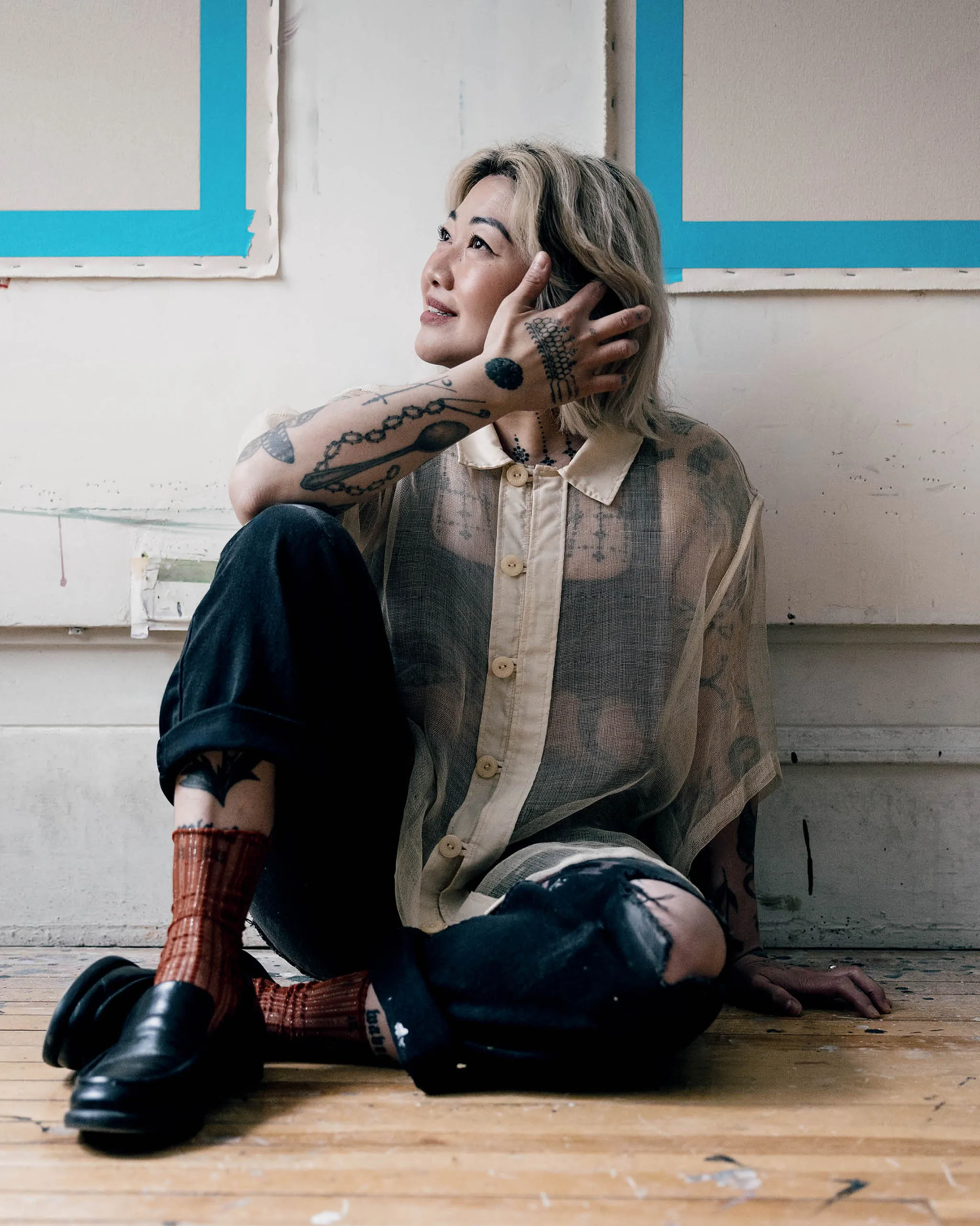 Person with blonde hair and tattoo sleeves sits on a wooden floor, resting head on their hand and gazing upward.