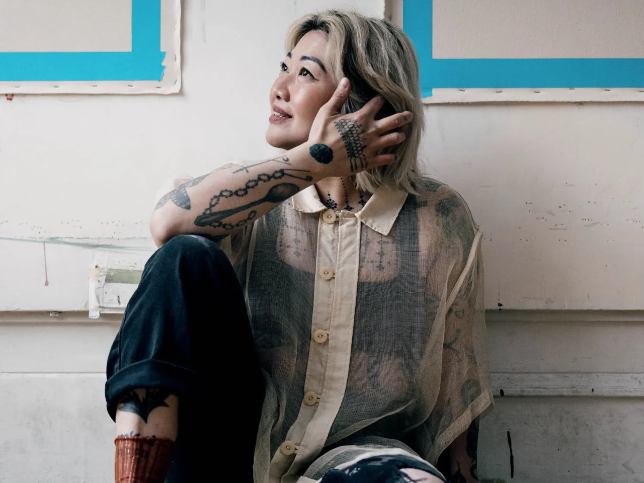 Woman with blond hair and multiple tattoos rests her head on her hand, looking upward in a worn room with blue tape on the wall.