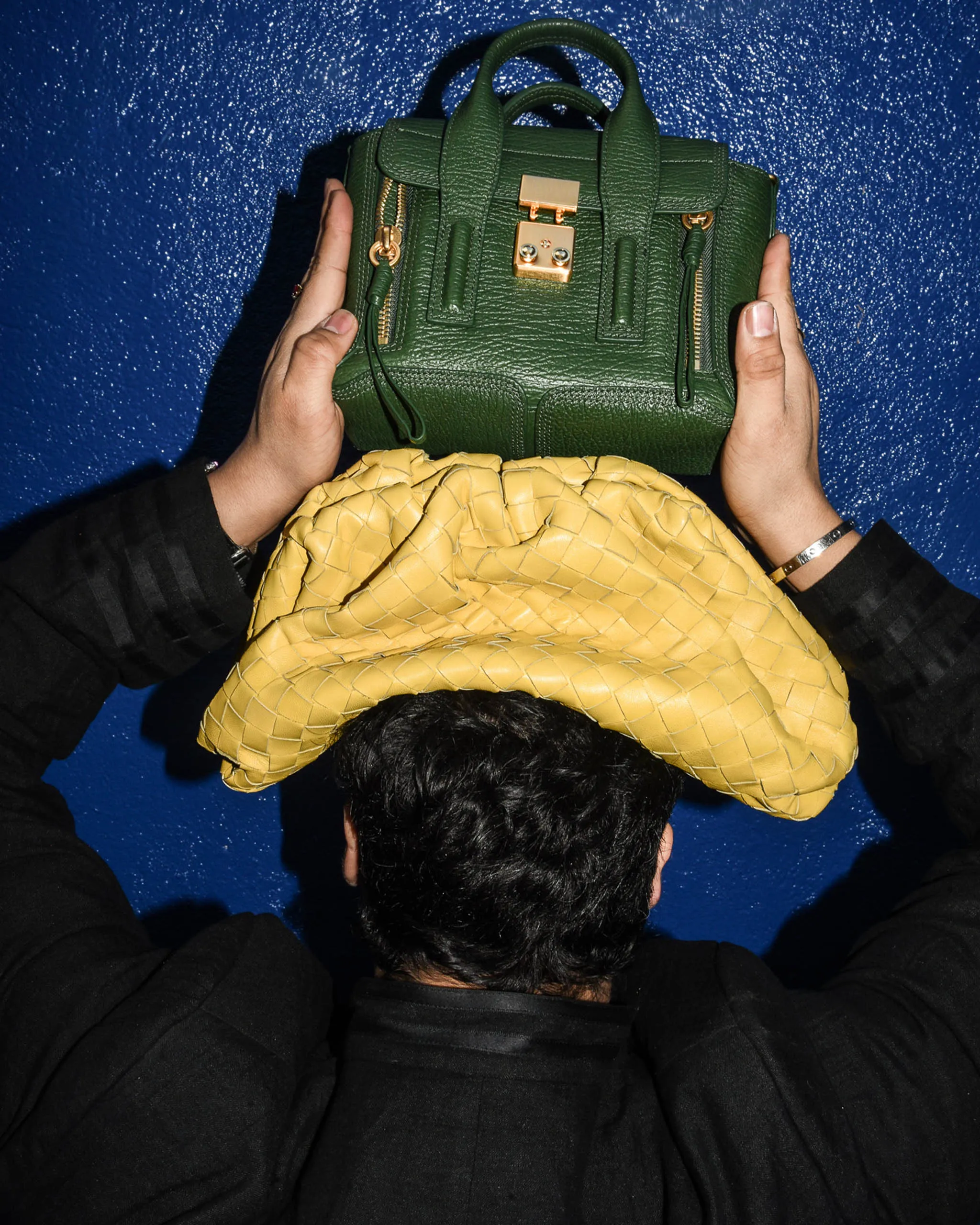Top-down view of a green leather handbag with gold hardware being held by a person in black, against a blue textured background.