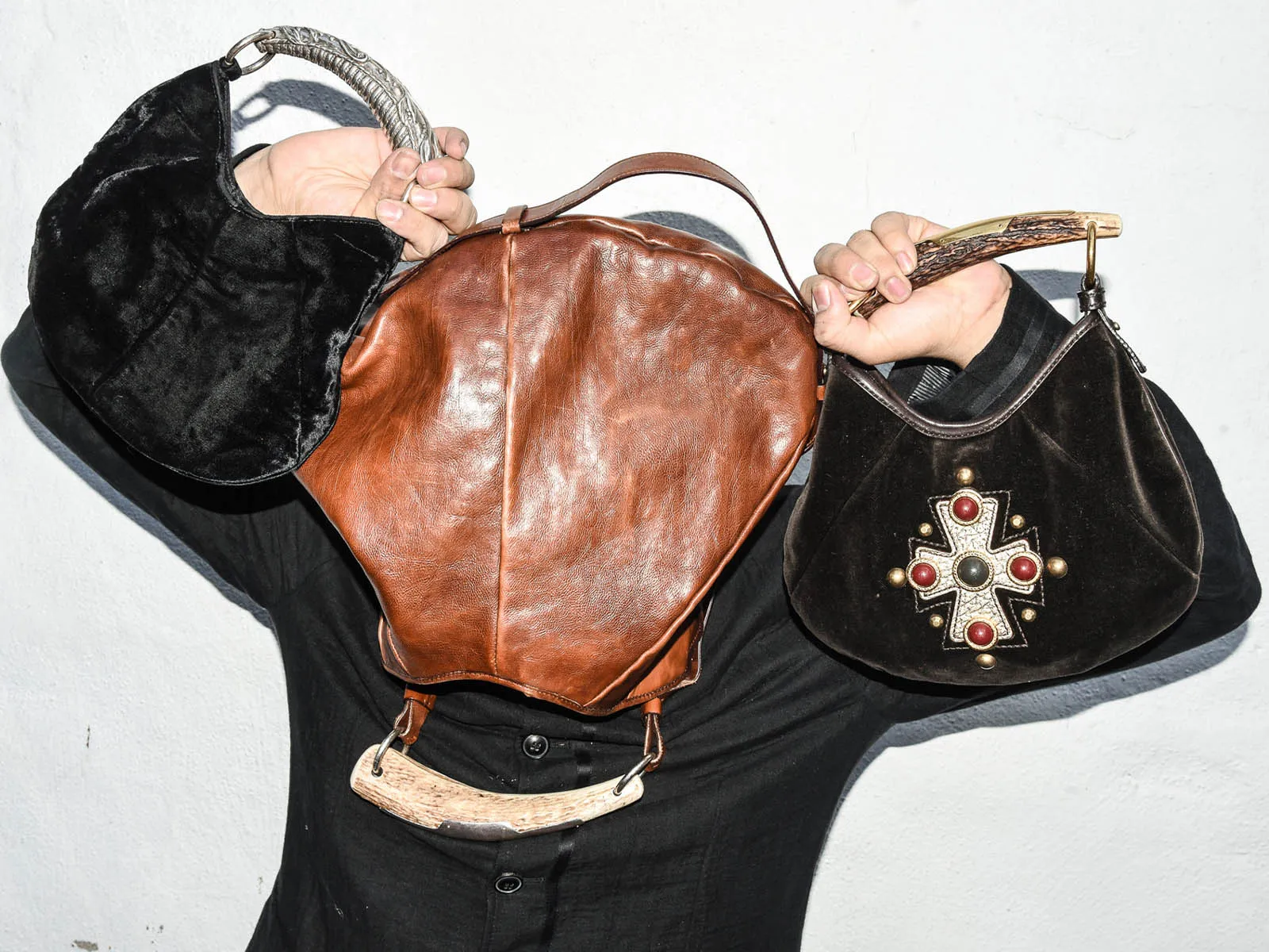 Person wearing black clothing holds up three vintage handbags: a brown leather backpack in the center and two black velvet bags with metal embellishments against a white wall.