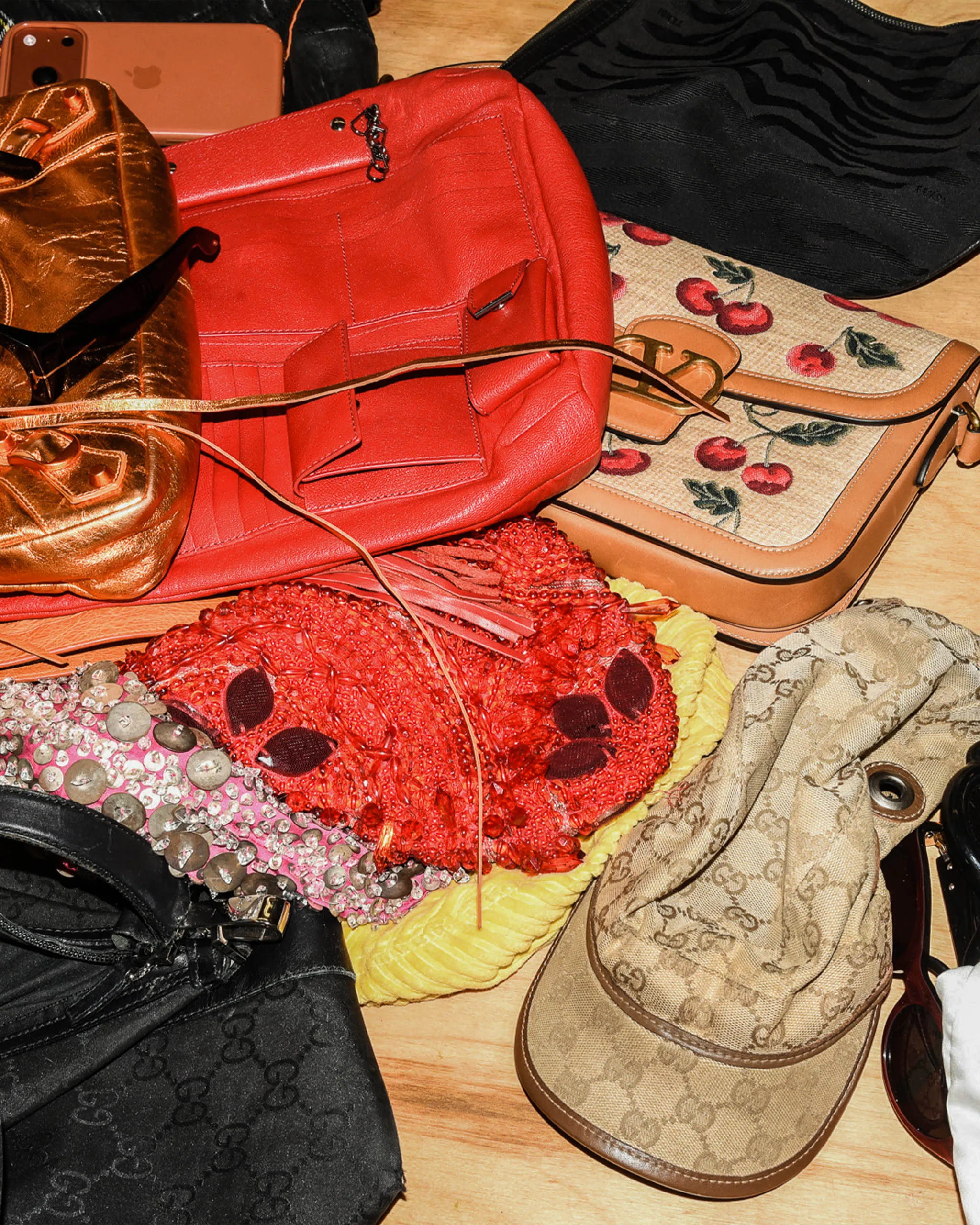 Red leather handbag with a front pocket sits on a tan Gucci monogram purse; a sparkling pink-red fabric drapes nearby on a wooden floor.