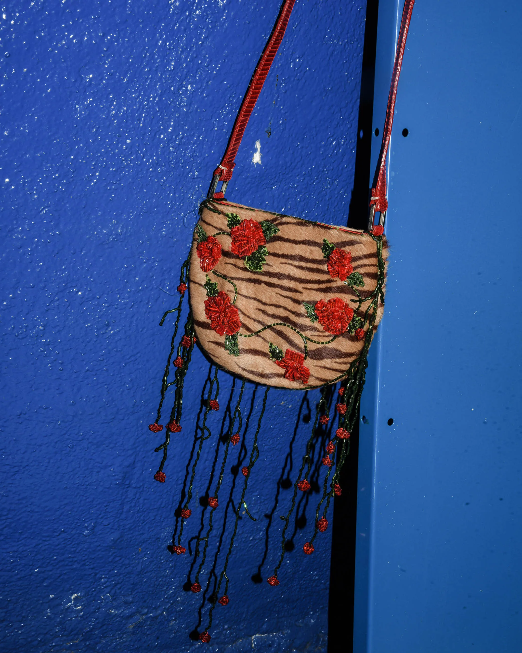Embroidered tiger-striped handbag with red flowers hanging from red straps against a blue wall backdrop.