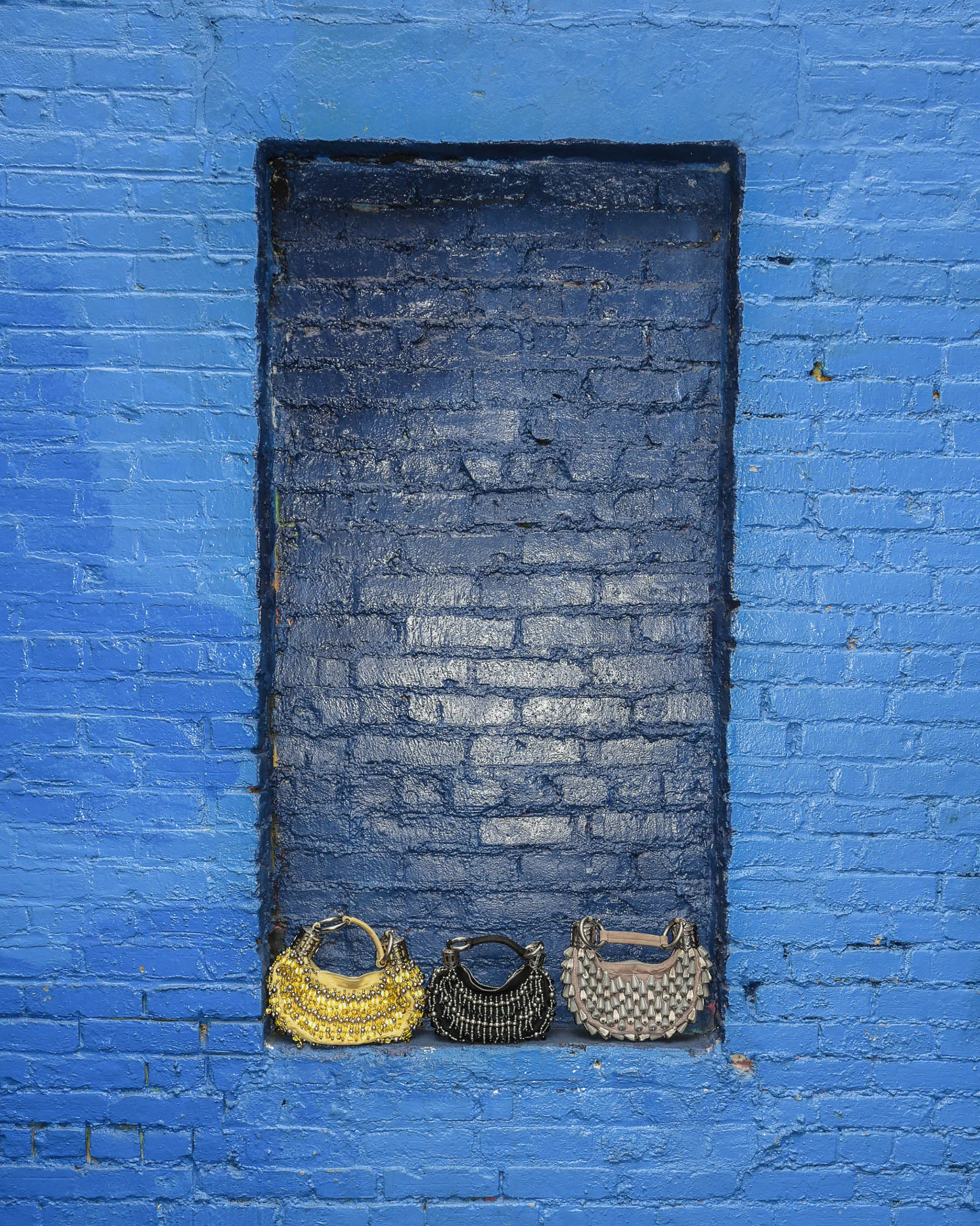 Blue brick wall with a dark rectangular recessed opening and three beaded handbags hanging along the bottom: gold, black, and silver.