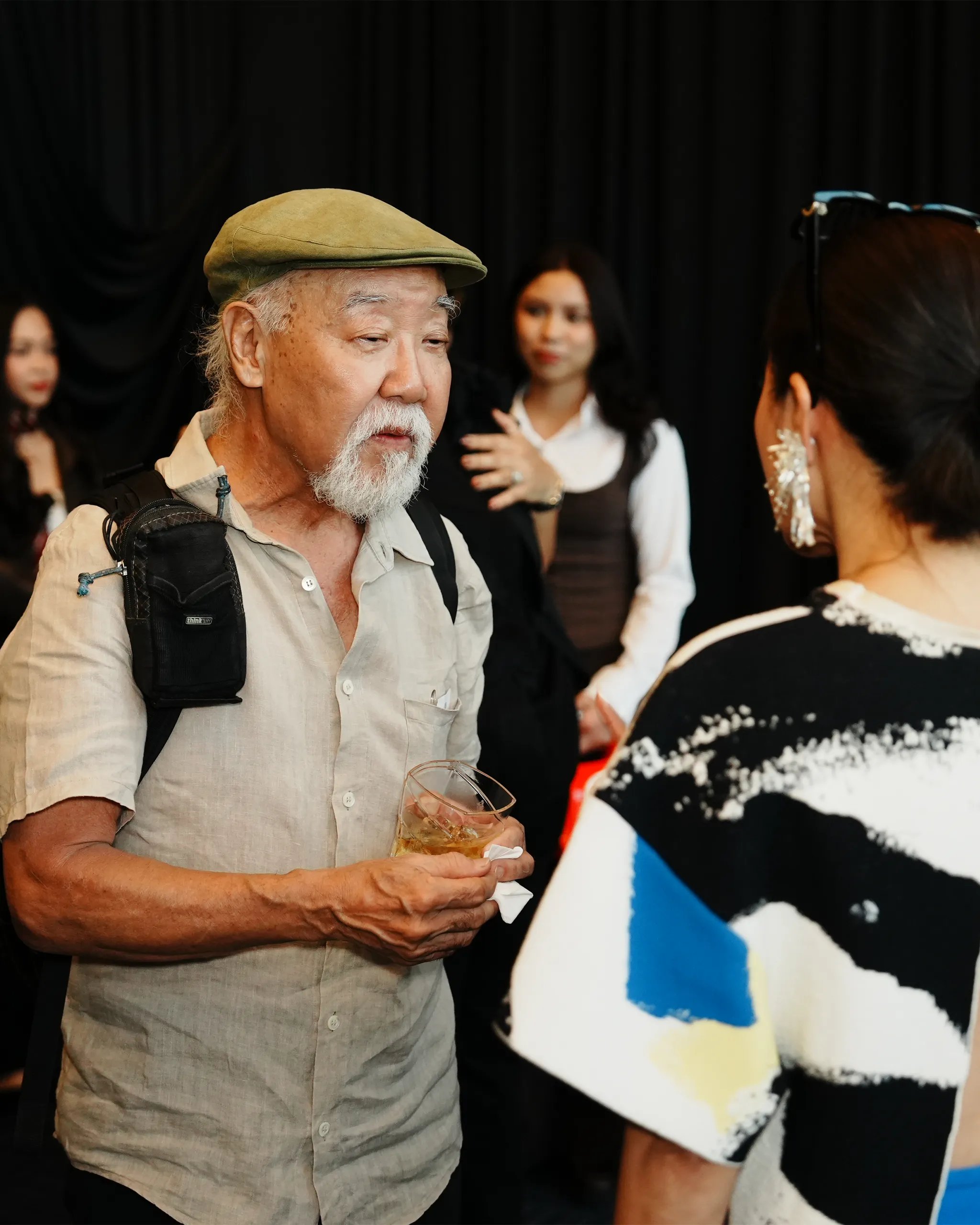 Older man with a beige cap and white beard holds a drink while talking to a woman in a bright patterned sweater at a social event in a dim room.