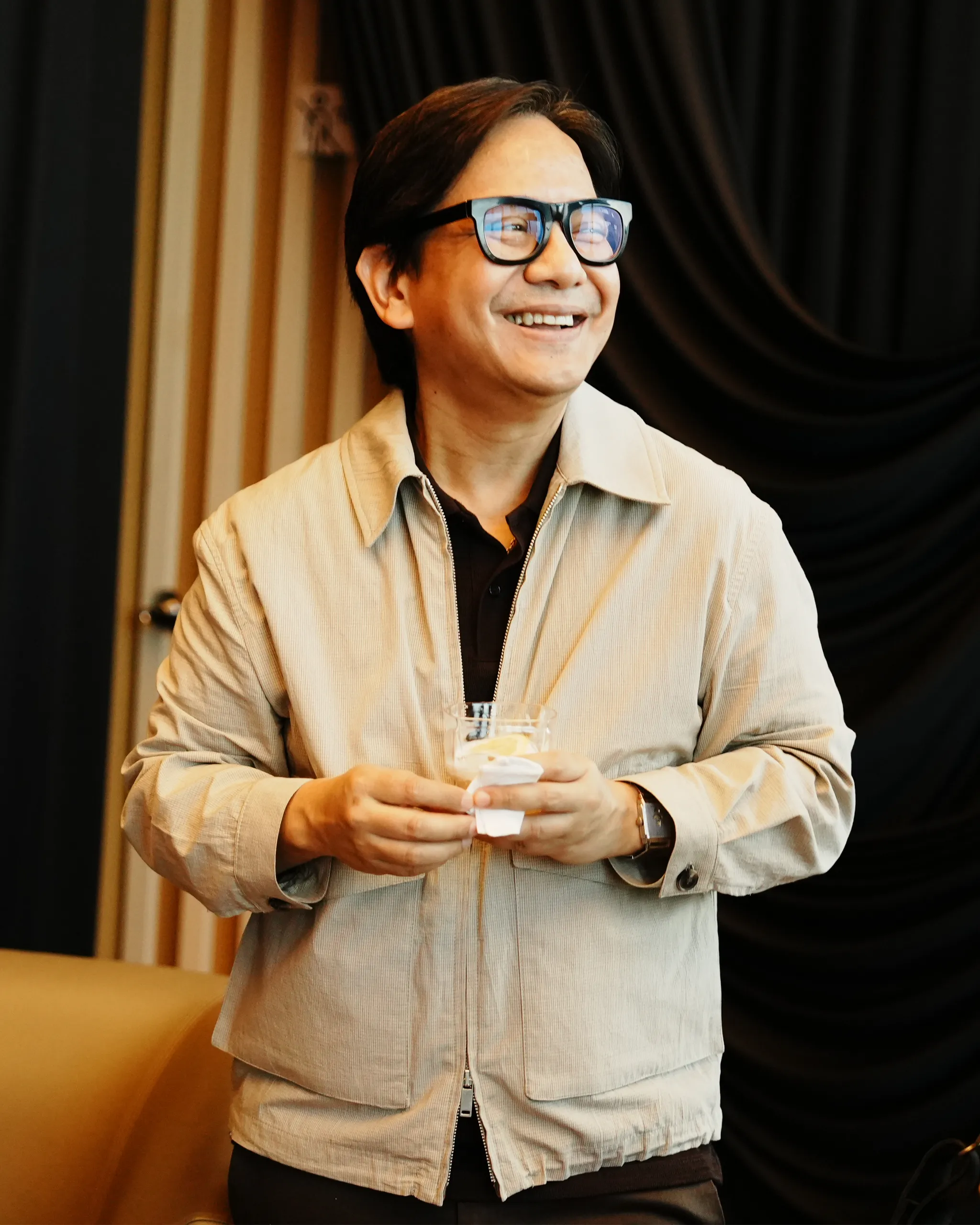 Smiling man with dark hair and glasses in a beige jacket, holding a napkin in front of him indoors.