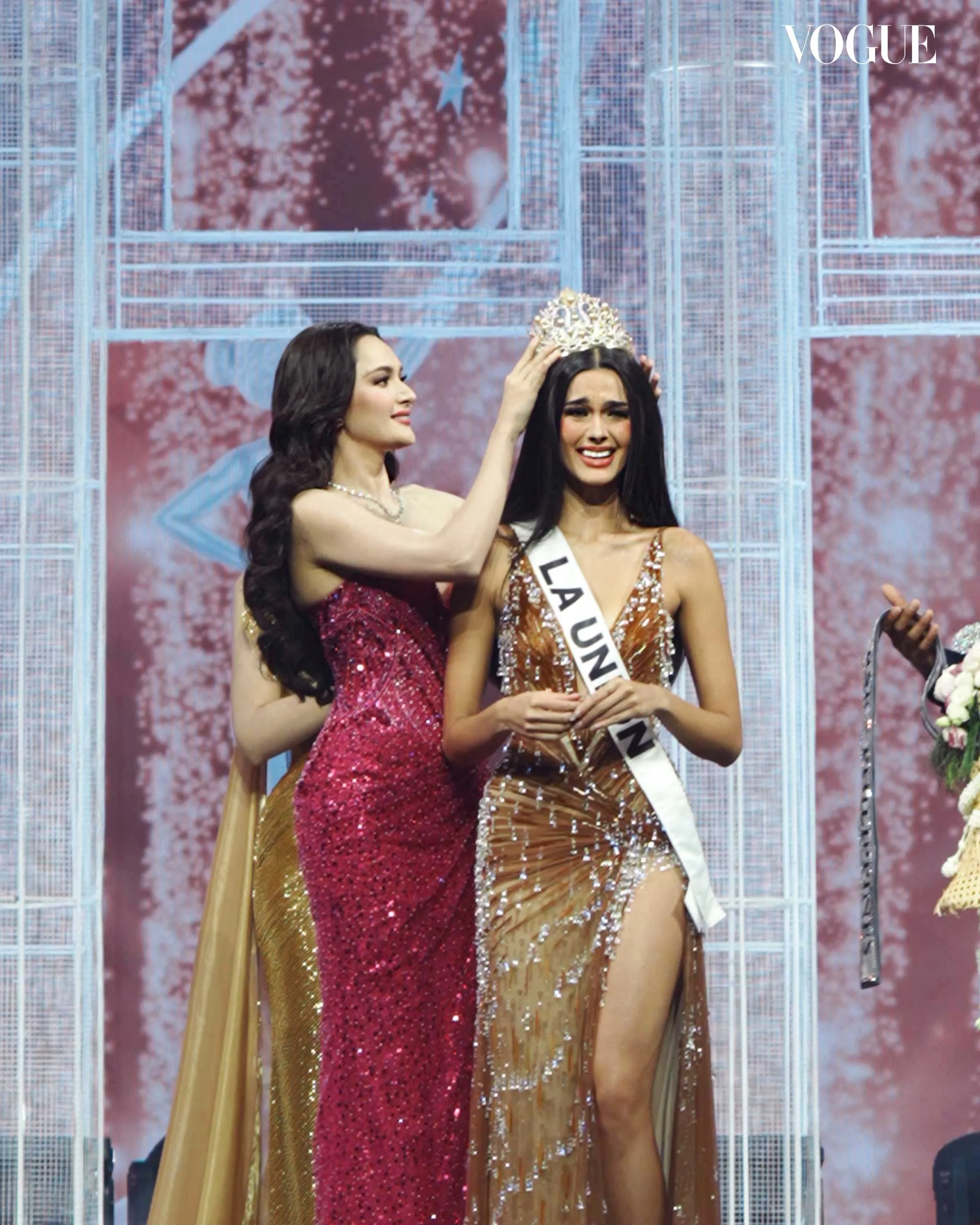 A contestant crowns the winner on stage as they wear glittering evening gowns at a beauty pageant with a pink backdrop.