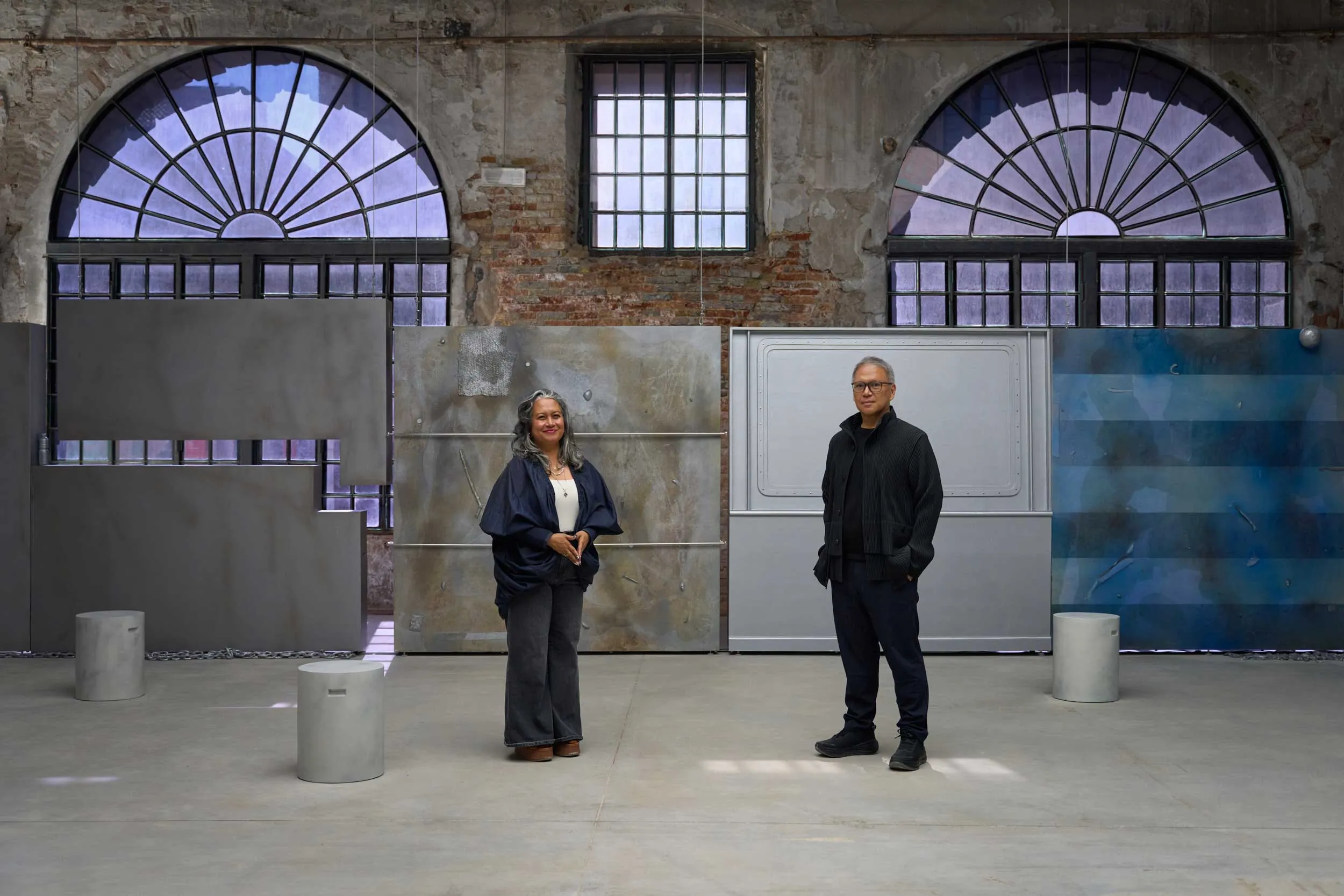 Two adults stand apart in an industrial, warehouse-like space with large arched windows and brick walls behind them.