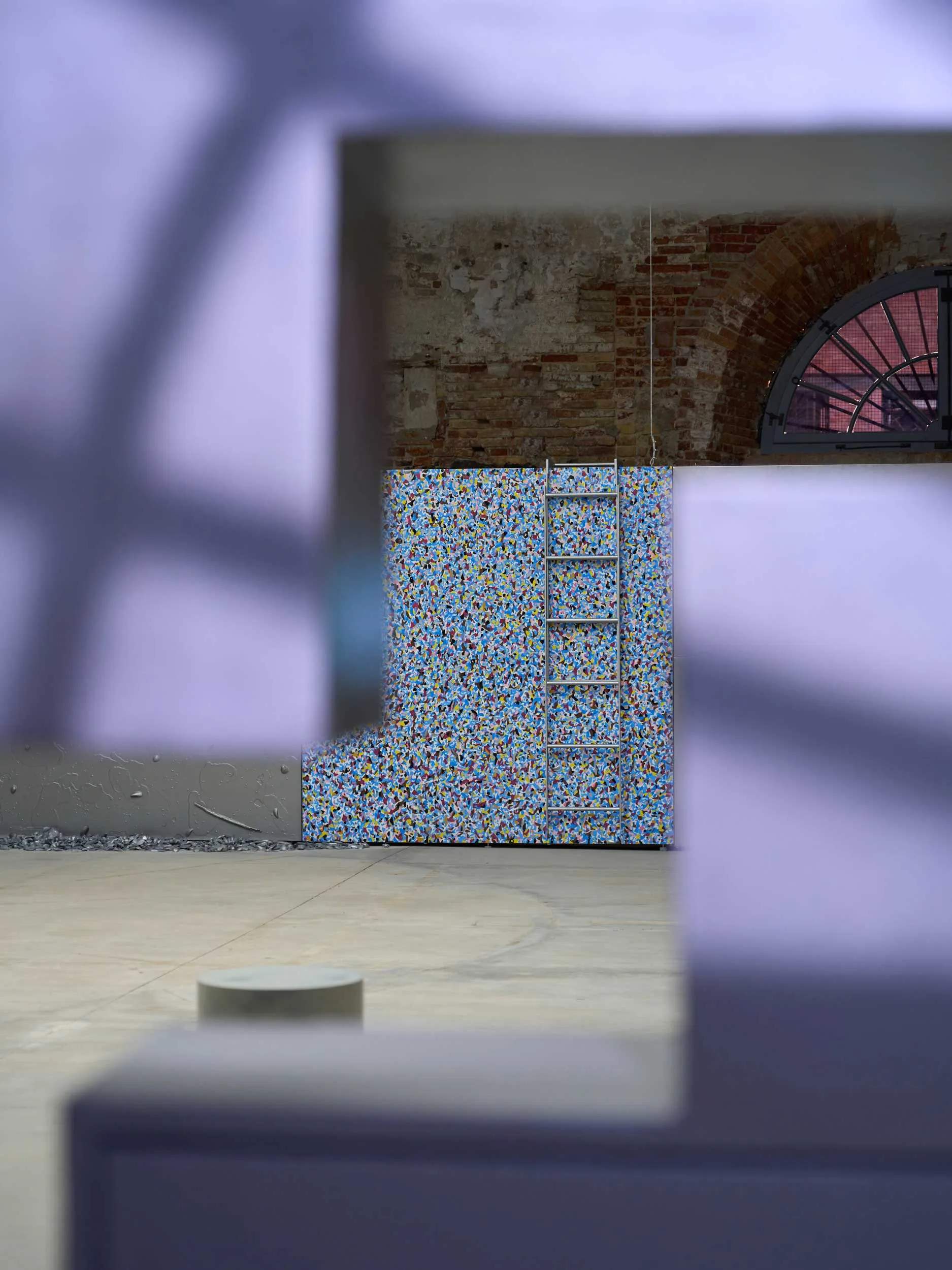 Interior with a colorful mosaic wall, a metal ladder leaning against it, and an exposed brick wall with a large arched window in the background; blurred foreground shapes frame the scene.