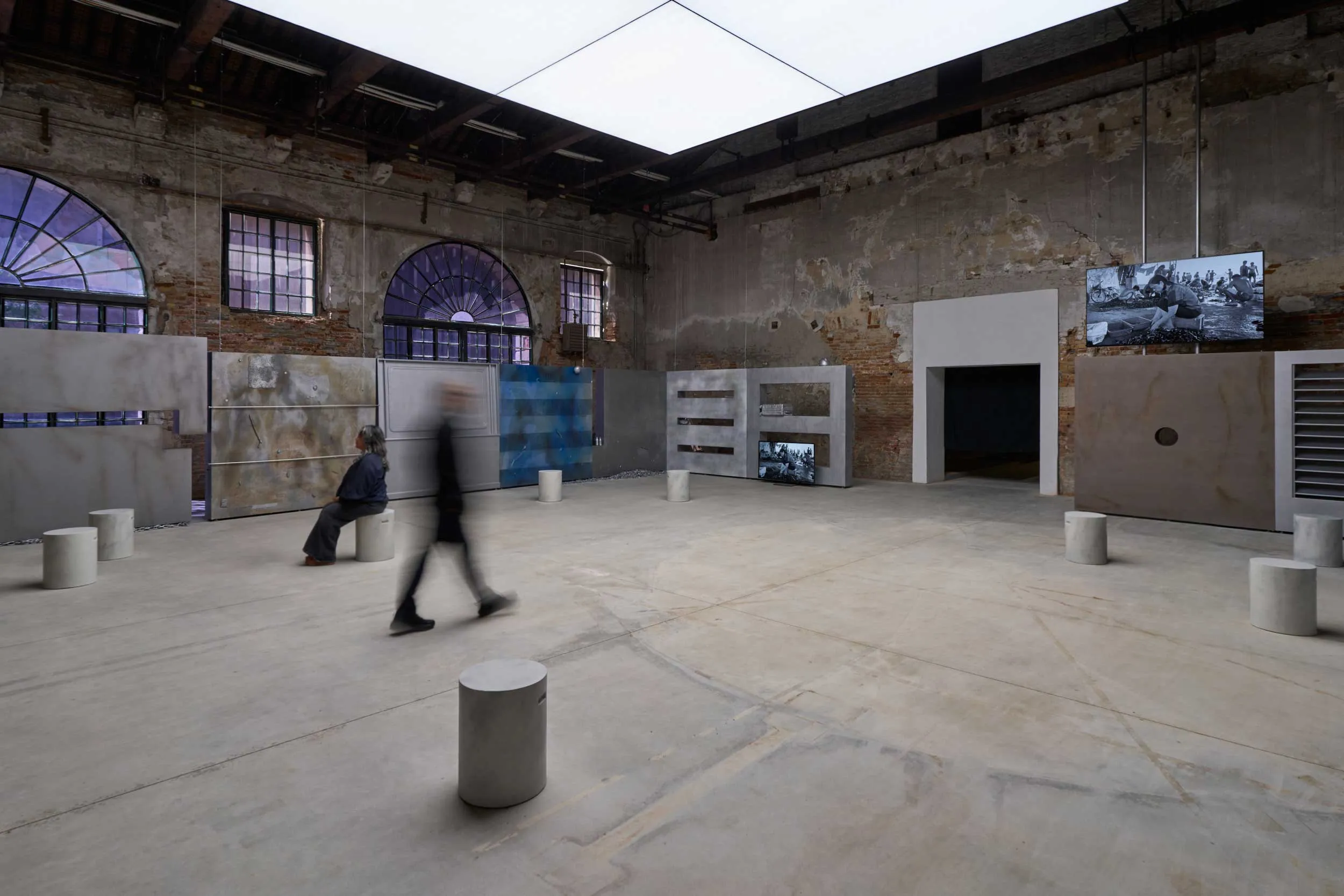 Industrial gallery space with brick walls, arched windows, and grey display panels; blurred visitors walk among cylindrical stools and large photos on the walls.