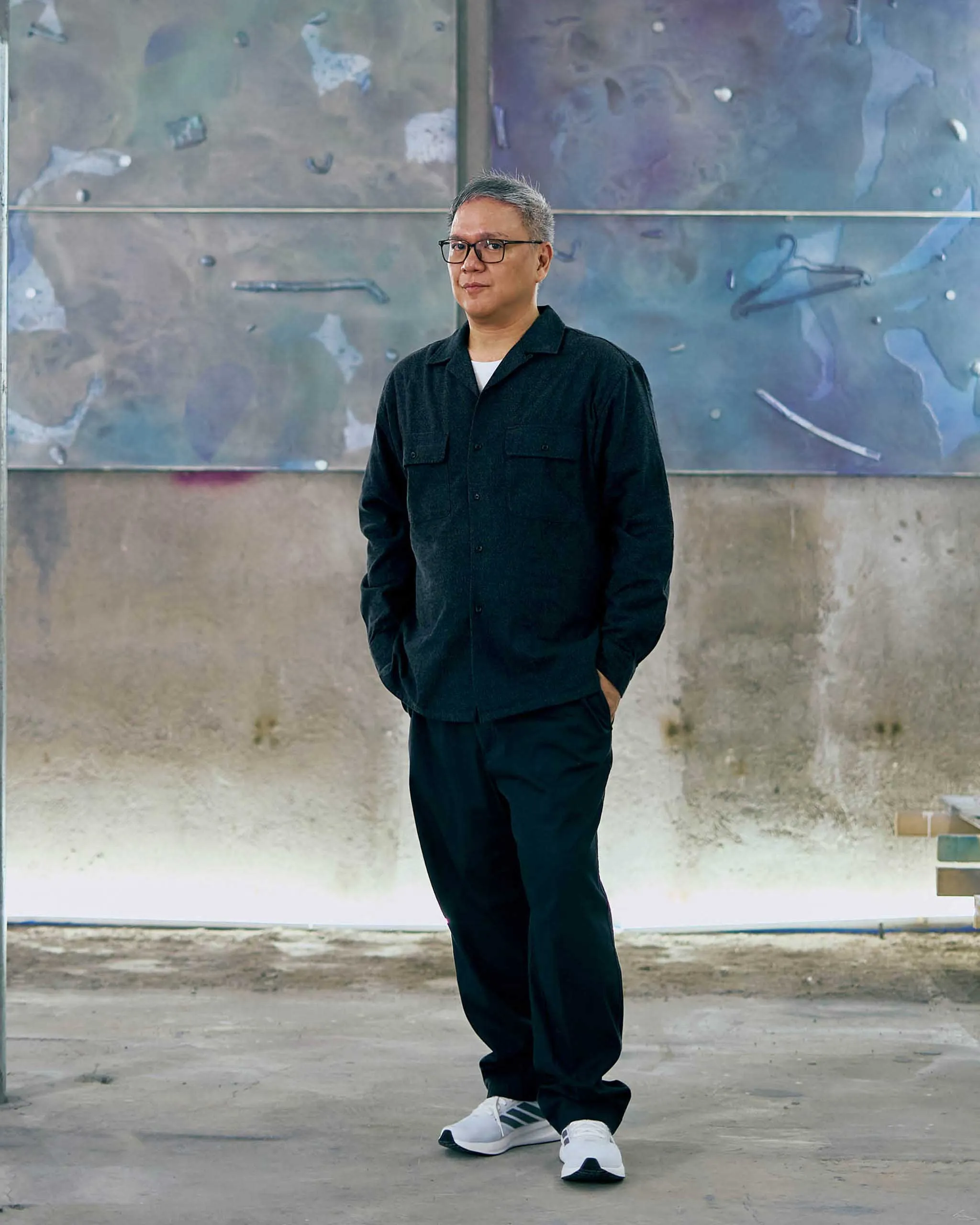 Man with short gray hair and glasses stands in front of a large, textured mural wearing a dark shirt and white sneakers.