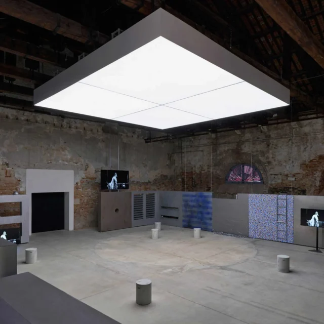 Industrial gallery with a large illuminated white cube hanging from the ceiling above display panels and brick walls.
