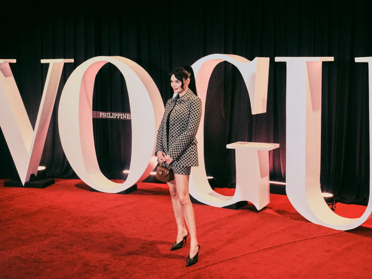A woman in a black-and-white checkered blazer posing on a red carpet in front of oversized white letters at an event backdrop.