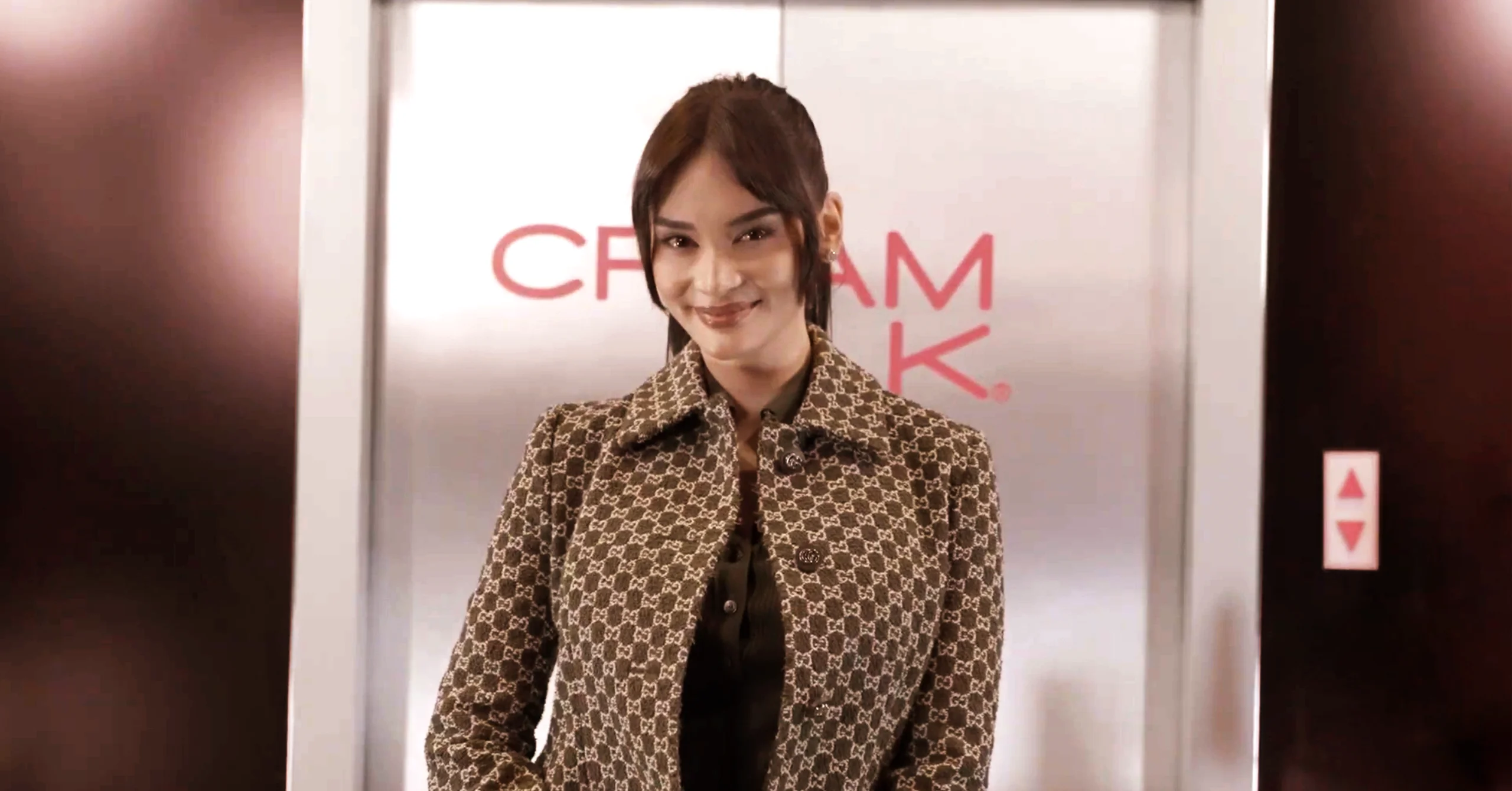 Smiling woman in a brown patterned coat stands in front of elevator doors in a lobby.