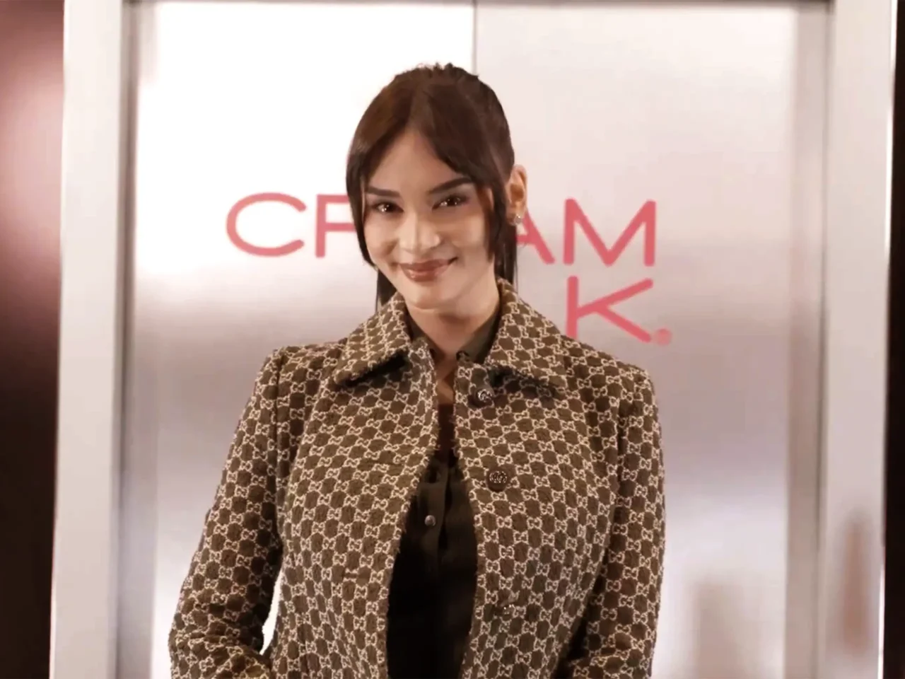 Smiling woman in a brown patterned coat stands in front of elevator doors in a lobby.
