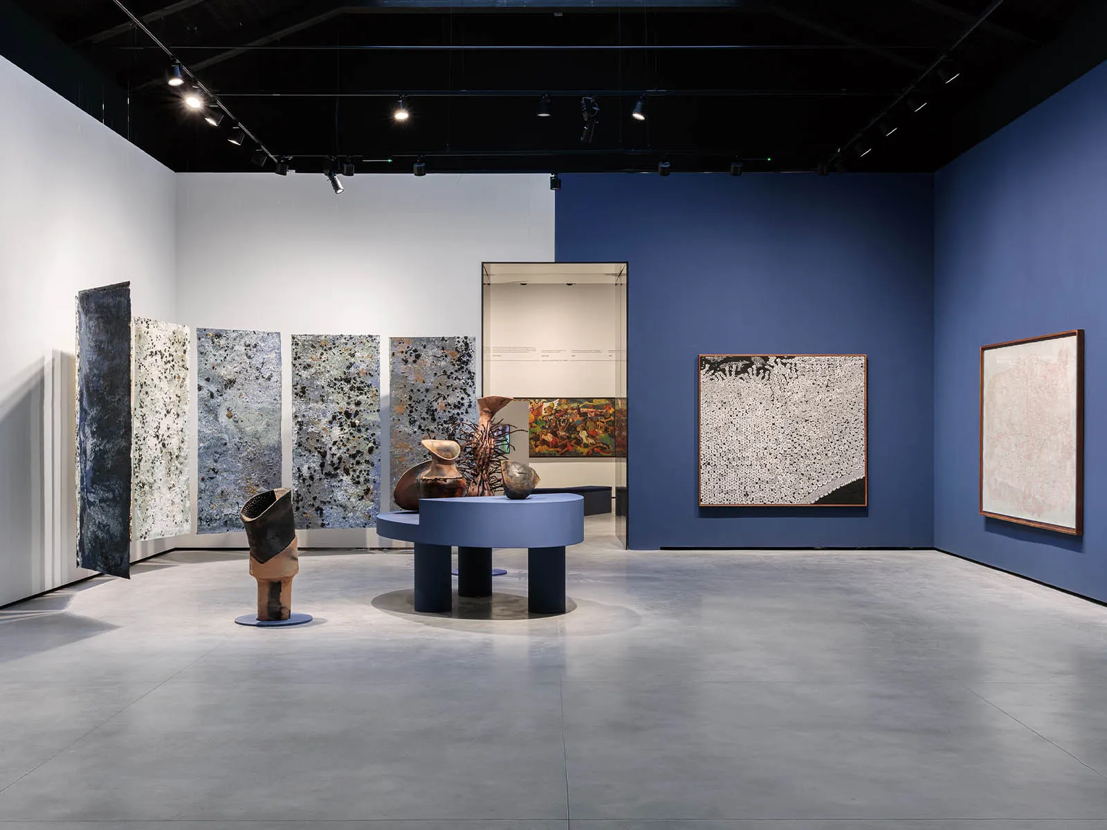 Art gallery interior with blue walls, textured vertical panels, and ceramic sculptures on a circular blue pedestal center stage.