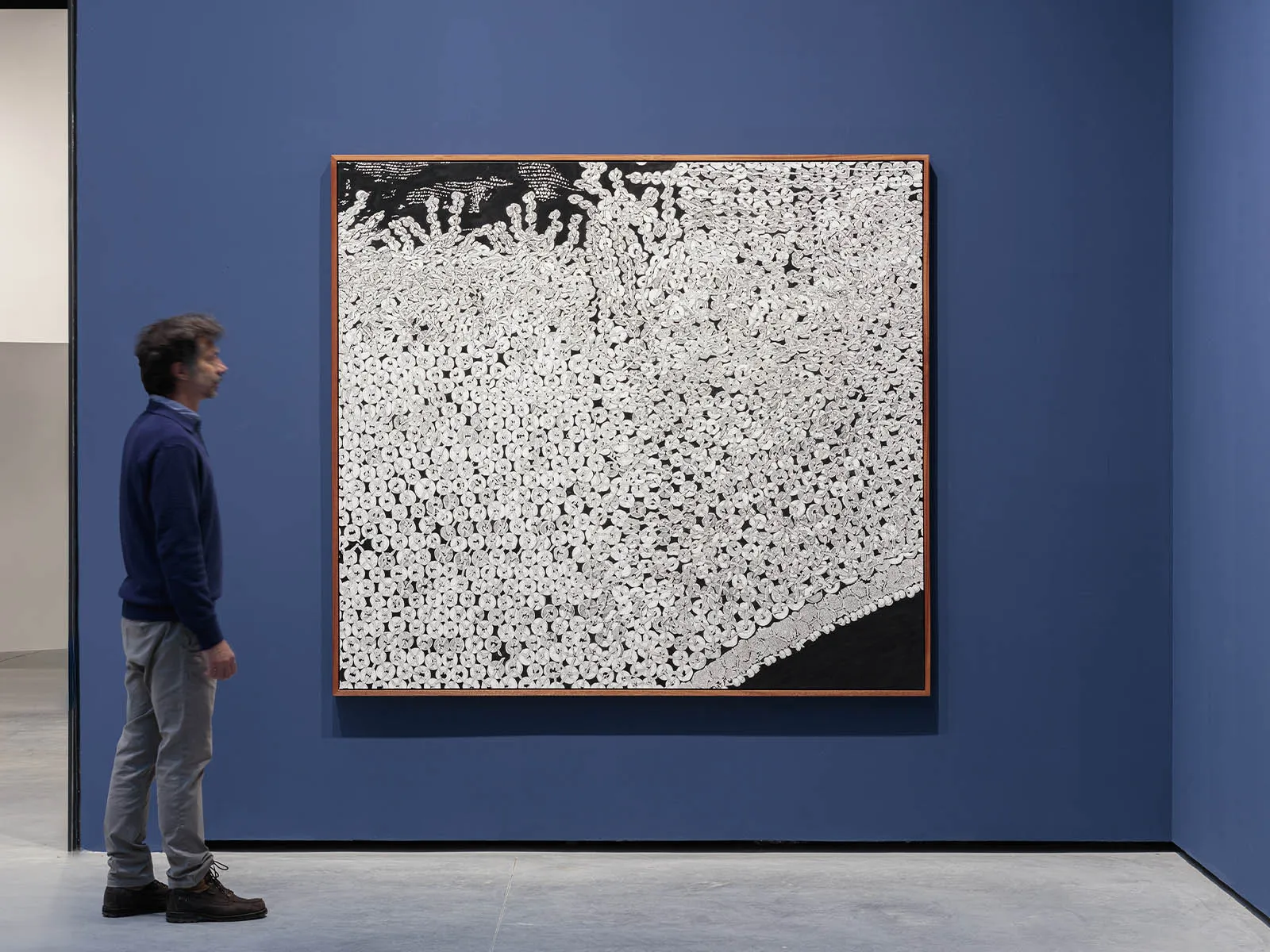 Man in a blue sweater and gray pants stands beside a large black-and-white abstract painting in a blue-walled gallery.