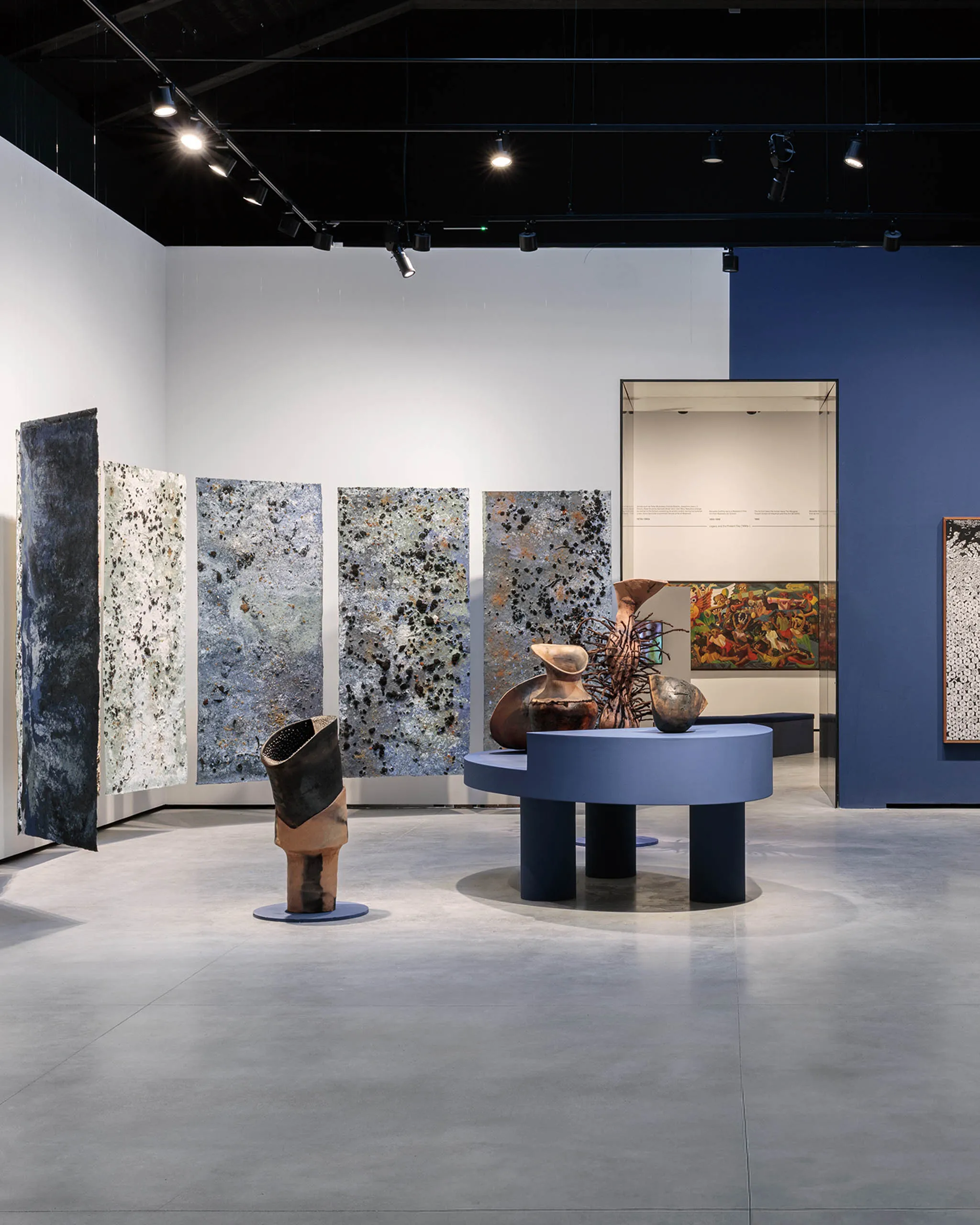 Art gallery interior with blue walls, tall speckled panels, and round blue pedestals displaying ceramic sculptures under track lighting.