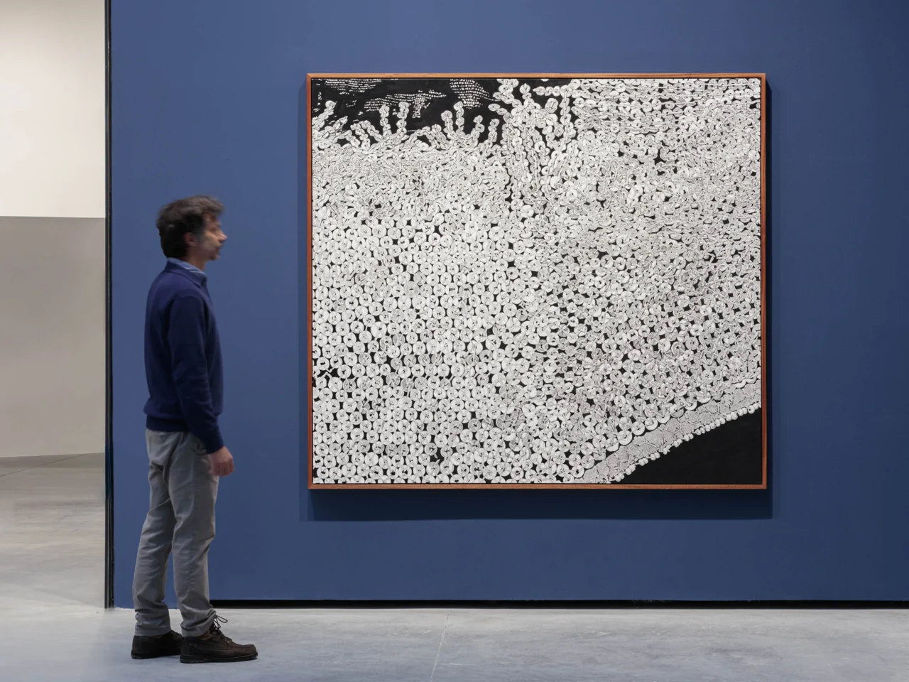 Blue-walled gallery with a doorway on the left revealing a patchwork mosaic of fabric-like panels and textures inside the next room; on the right, black-and-white framed drawings arranged in two rows along the wall.