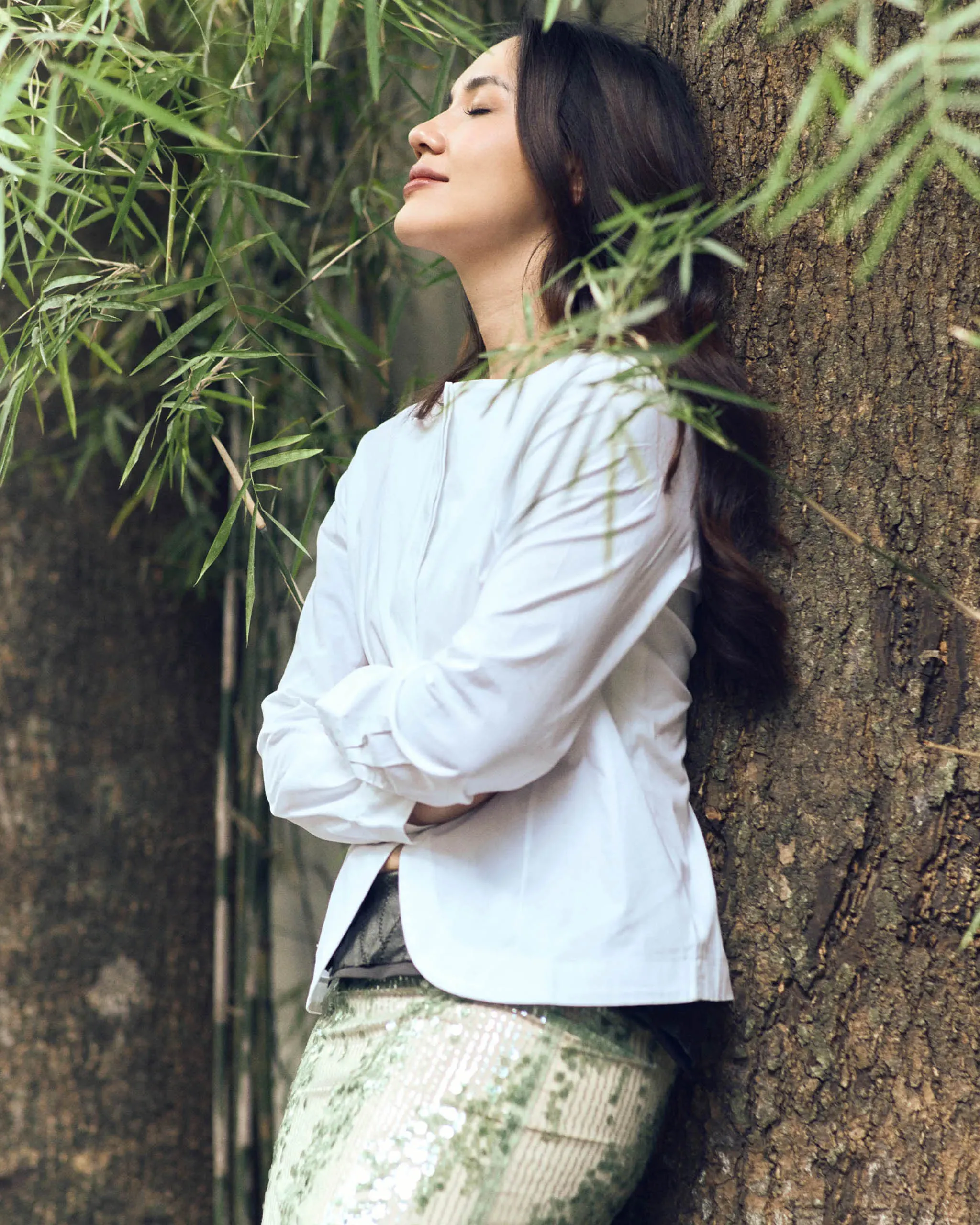 Woman leaning against a tree trunk with eyes closed, surrounded by bamboo leaves in a calm outdoor setting, wearing a light blue shirt.