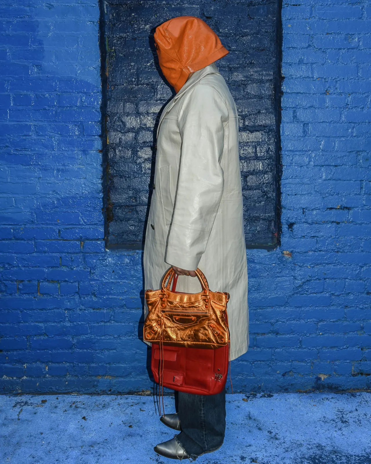 Person in a black robe grips a brown leather backpack with a curved wooden handle against a light background, palms visible near the bag’s strap.