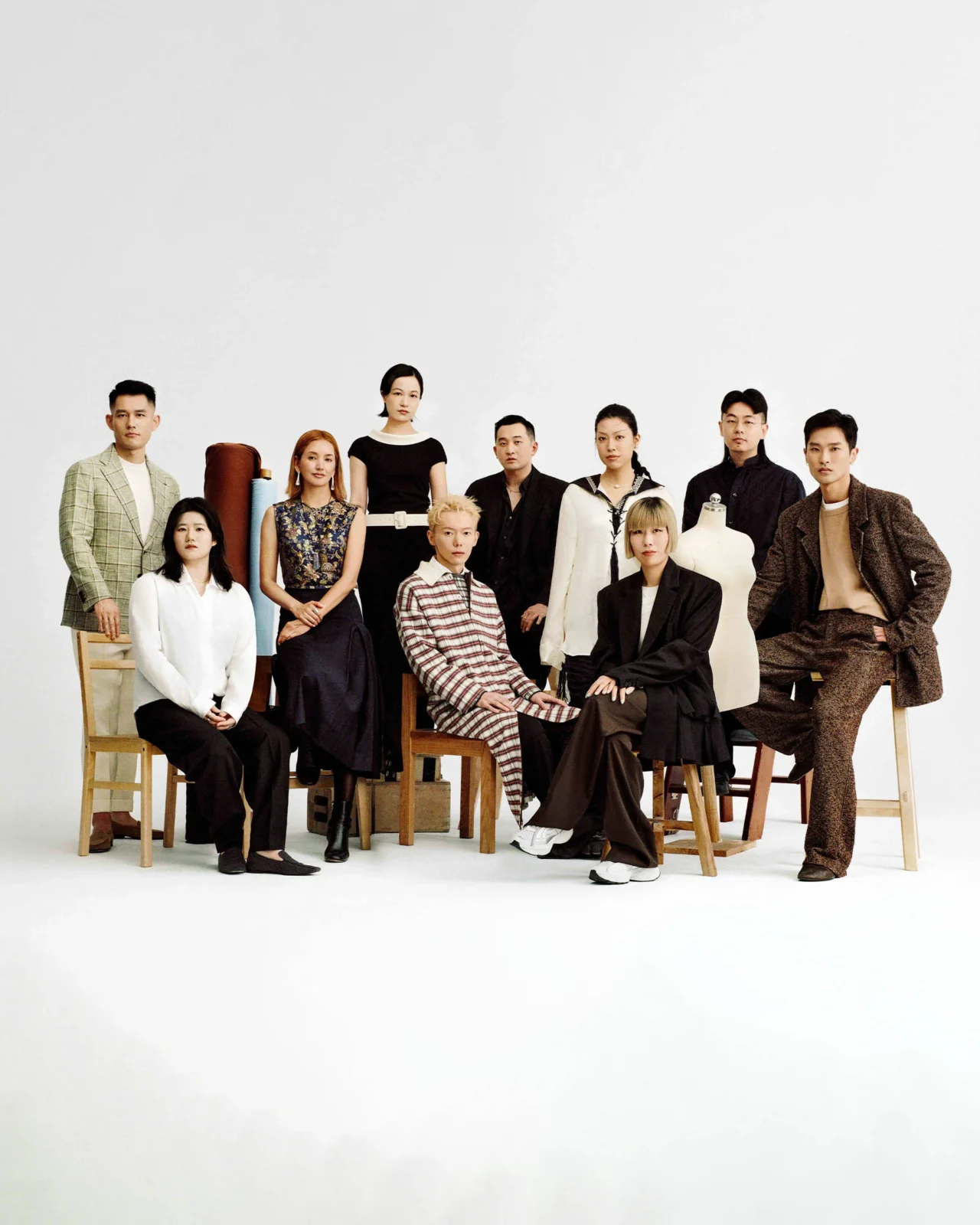 Group of ten fashion models posing in a studio against a white backdrop, wearing diverse outfits and sitting or standing with stools and mannequins nearby.