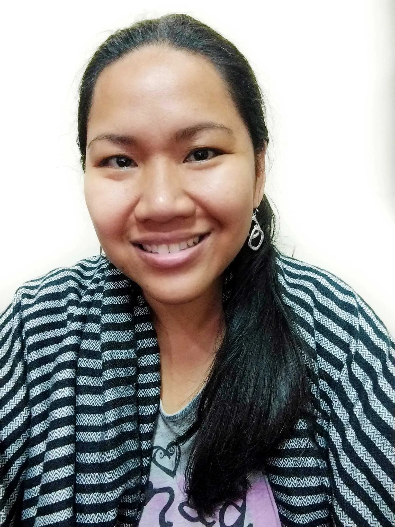 Close-up portrait of a smiling woman with long dark hair, wearing a black-and-white striped cardigan and silver earrings against a light background.