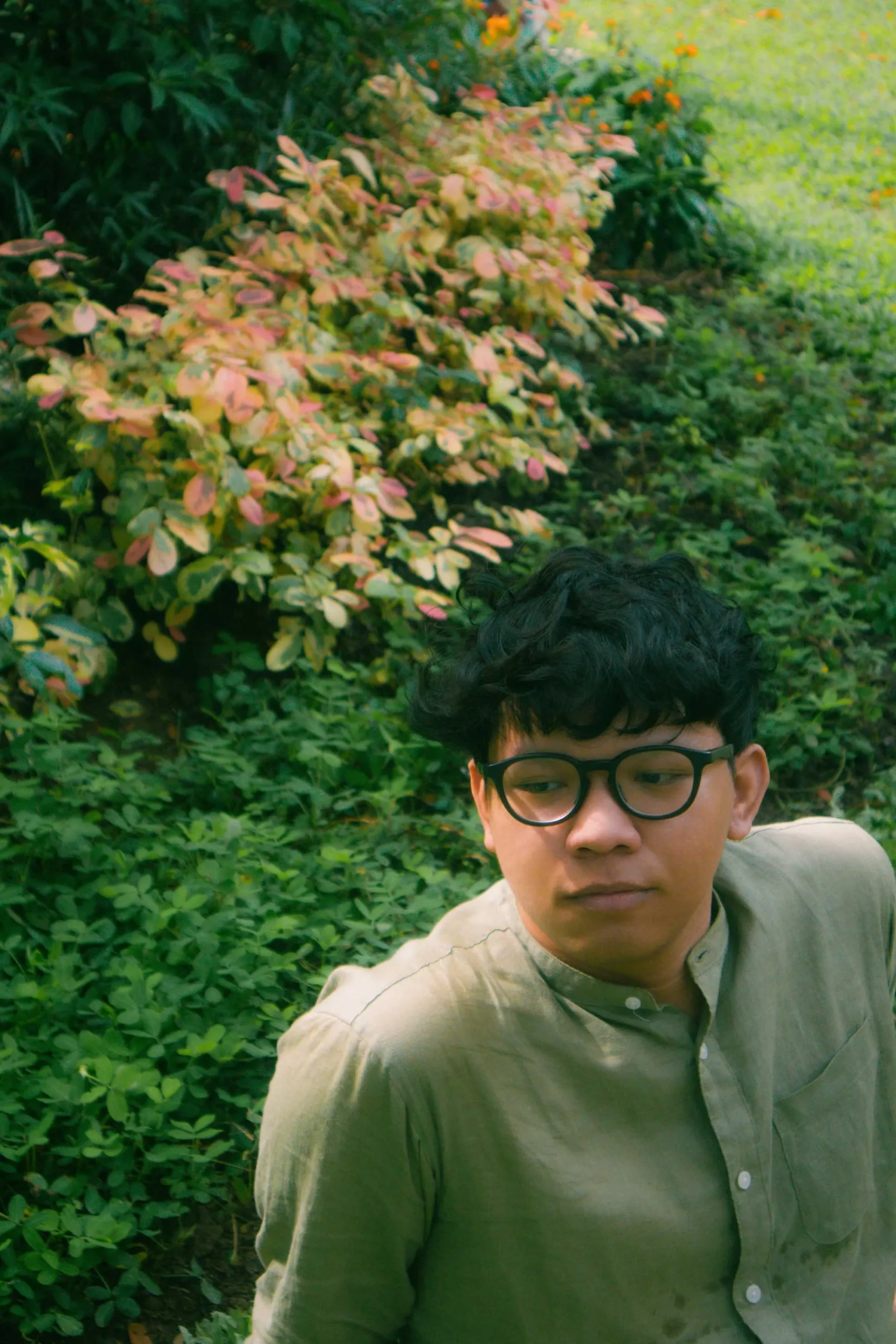 Young man wearing glasses in a garden with dense green foliage and pink-tinted leaves in the background.