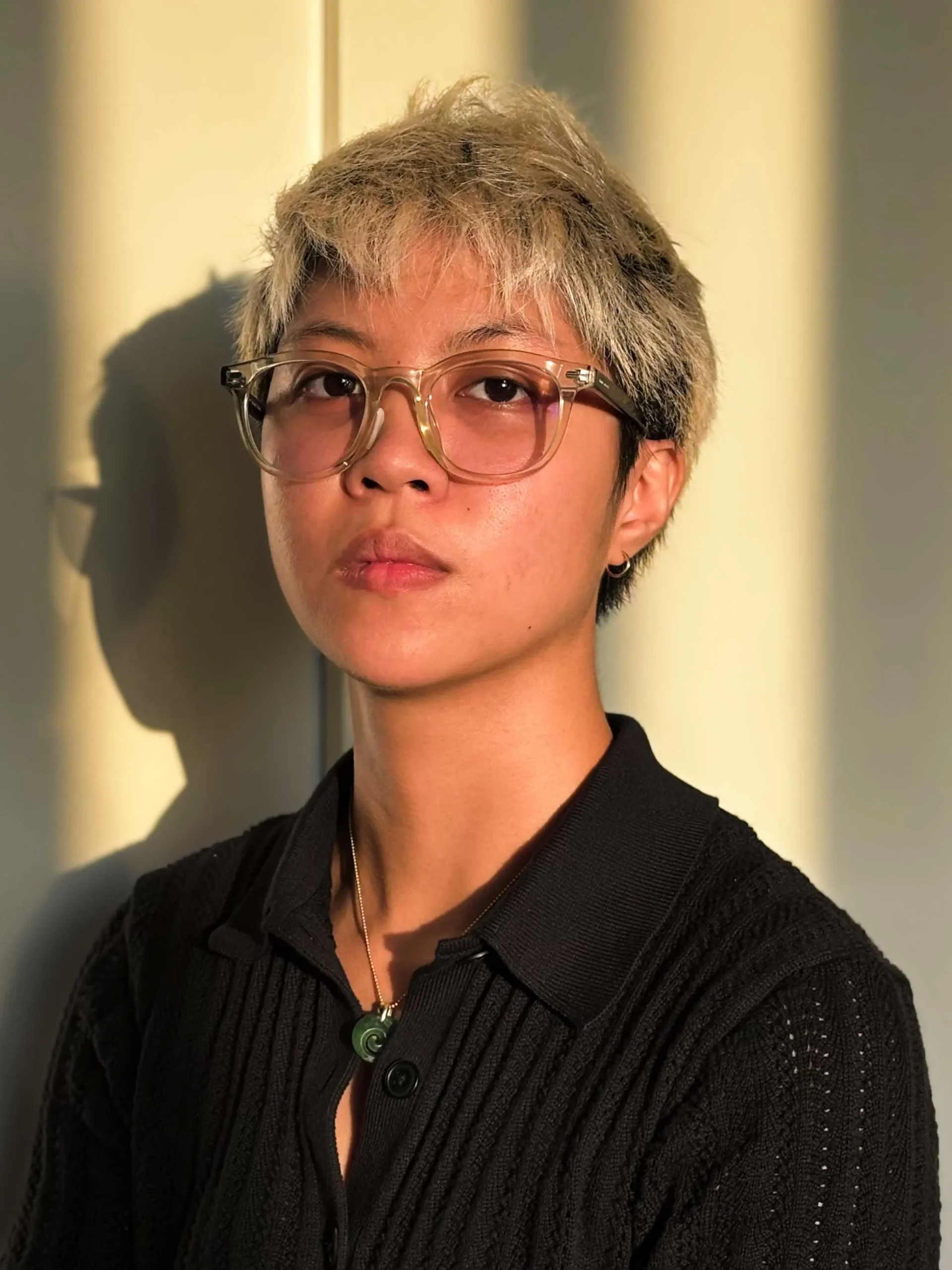 Person with short blond-streaked hair wearing clear-framed glasses and a black knit cardigan, looking slightly to the left in warm light.