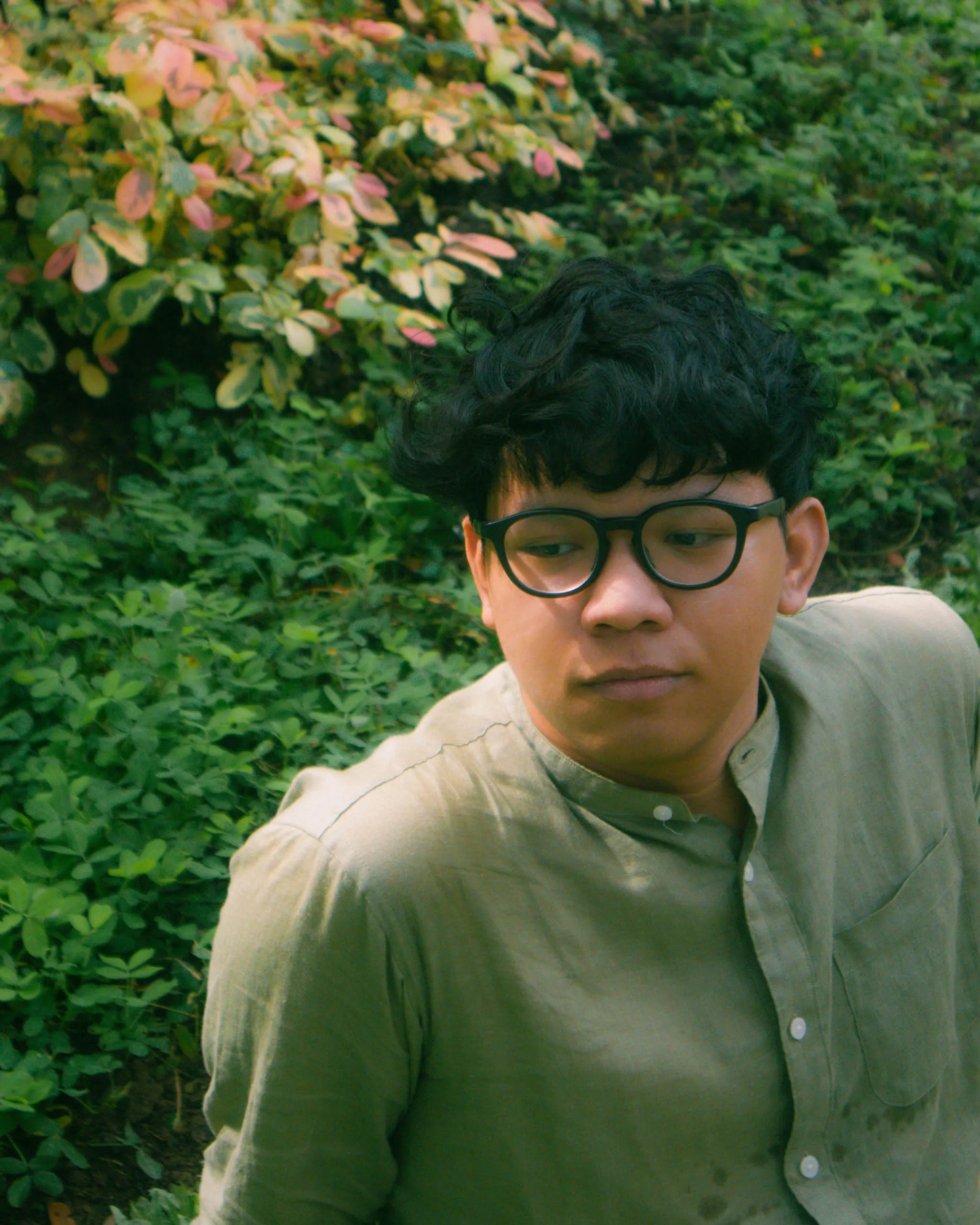 Young man with dark hair and glasses leaning outdoors among green bushes and leaves.