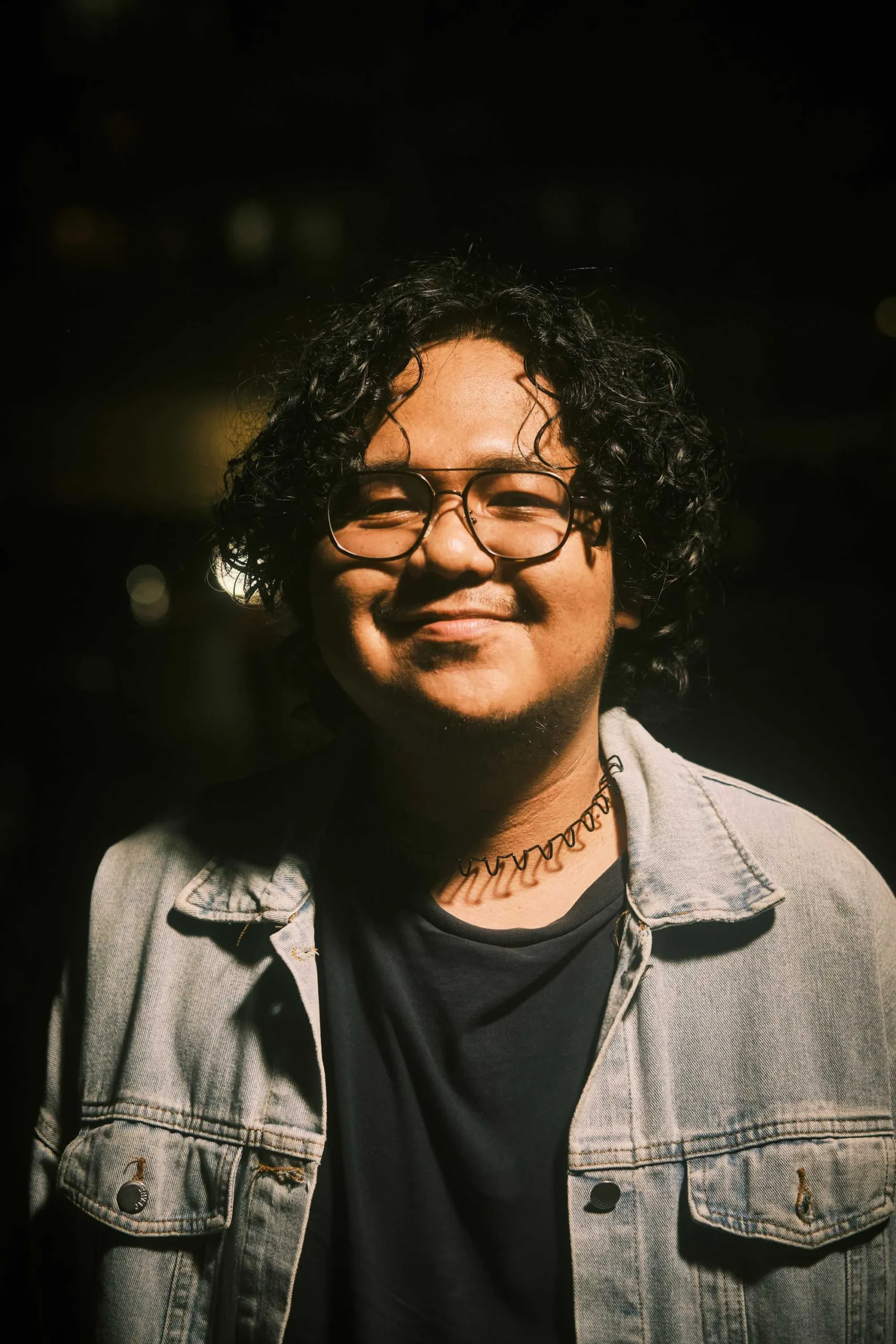 Smiling young man with curly hair and round glasses wearing a light denim jacket, photographed against a dark background at night.
