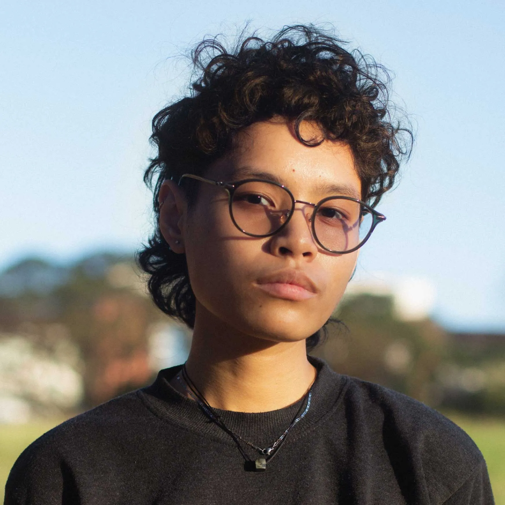 Young person with short curly hair and round glasses outdoors, wearing a black shirt and necklace under a clear sky.