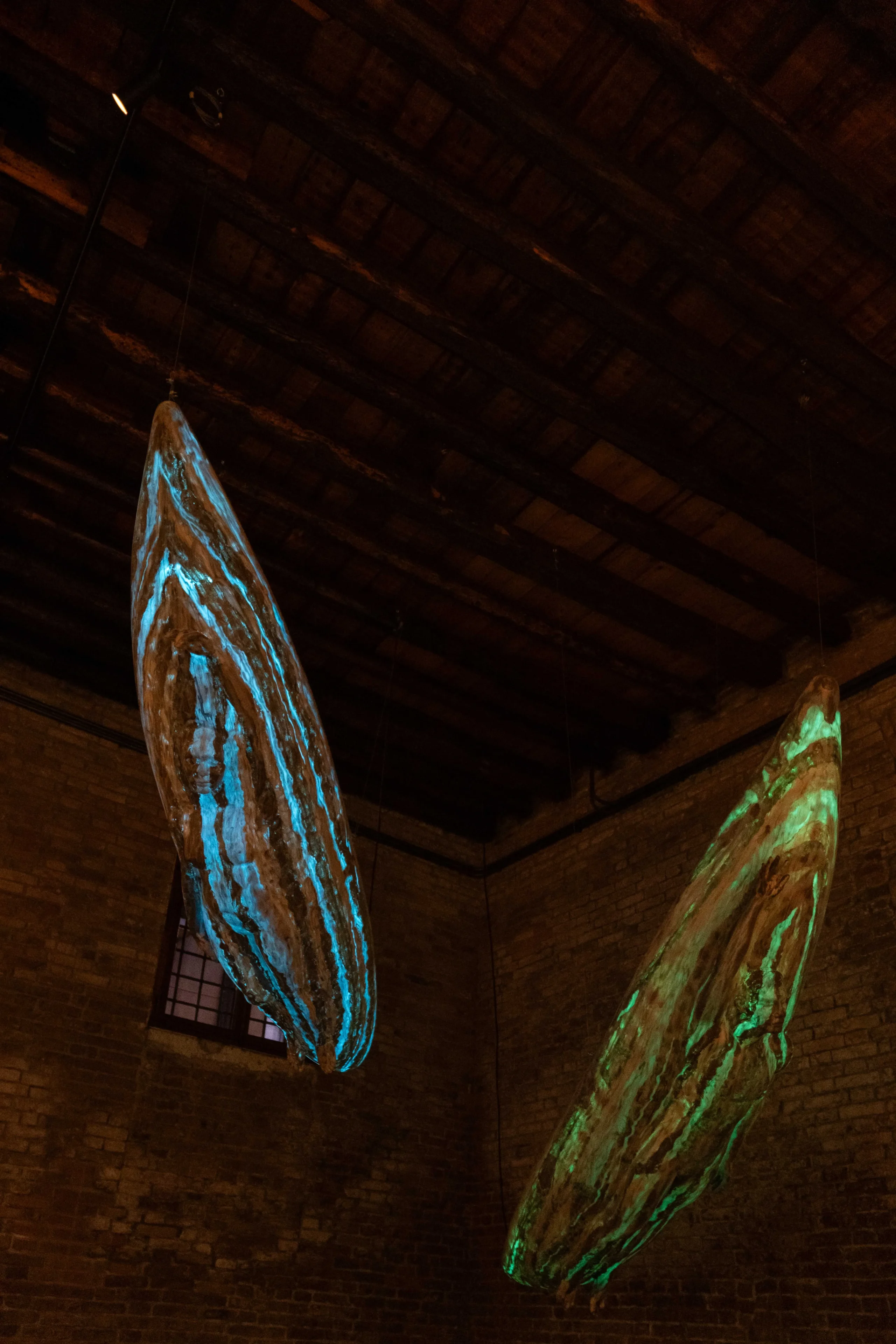 Hanging glass sculpture resembling a long blue-green blade against a dark brick ceiling interior.