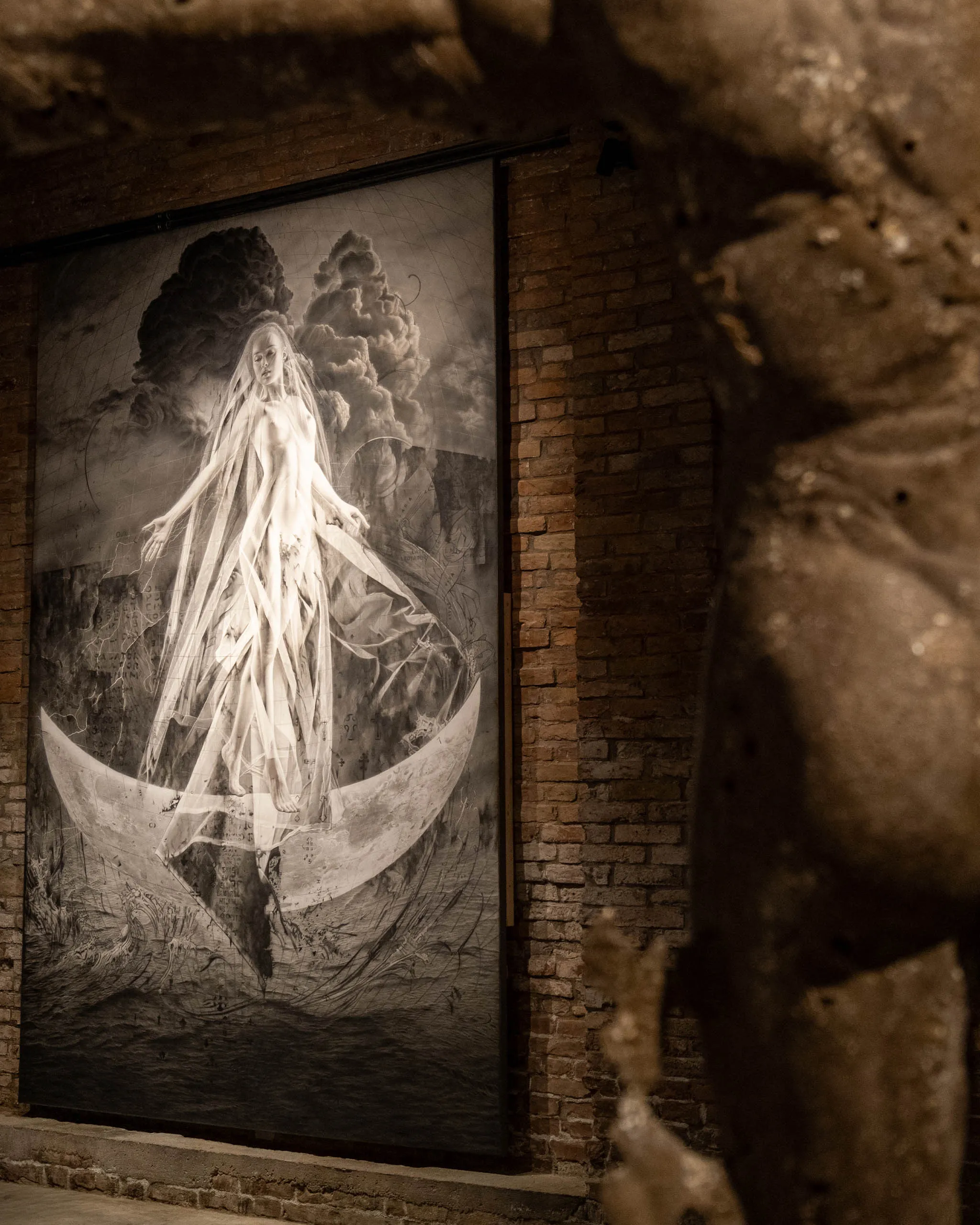 Black-and-white portrait of a veiled woman in a flowing gown, set against dramatic clouds on a brick wall in a dim space.