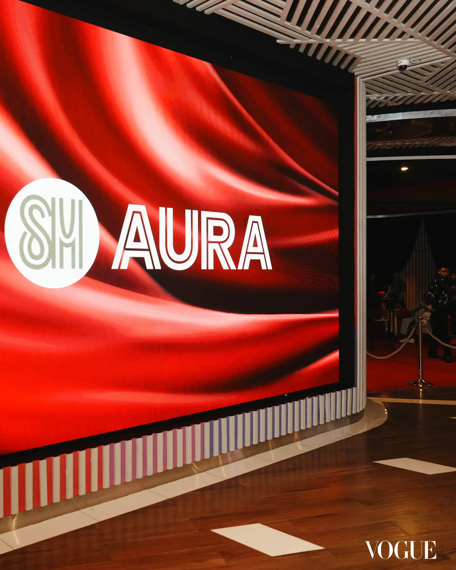 Large red curved backdrop with the word 'AURA' in white letters and a circular logo on the left; Vogue watermark in the bottom-right corner.