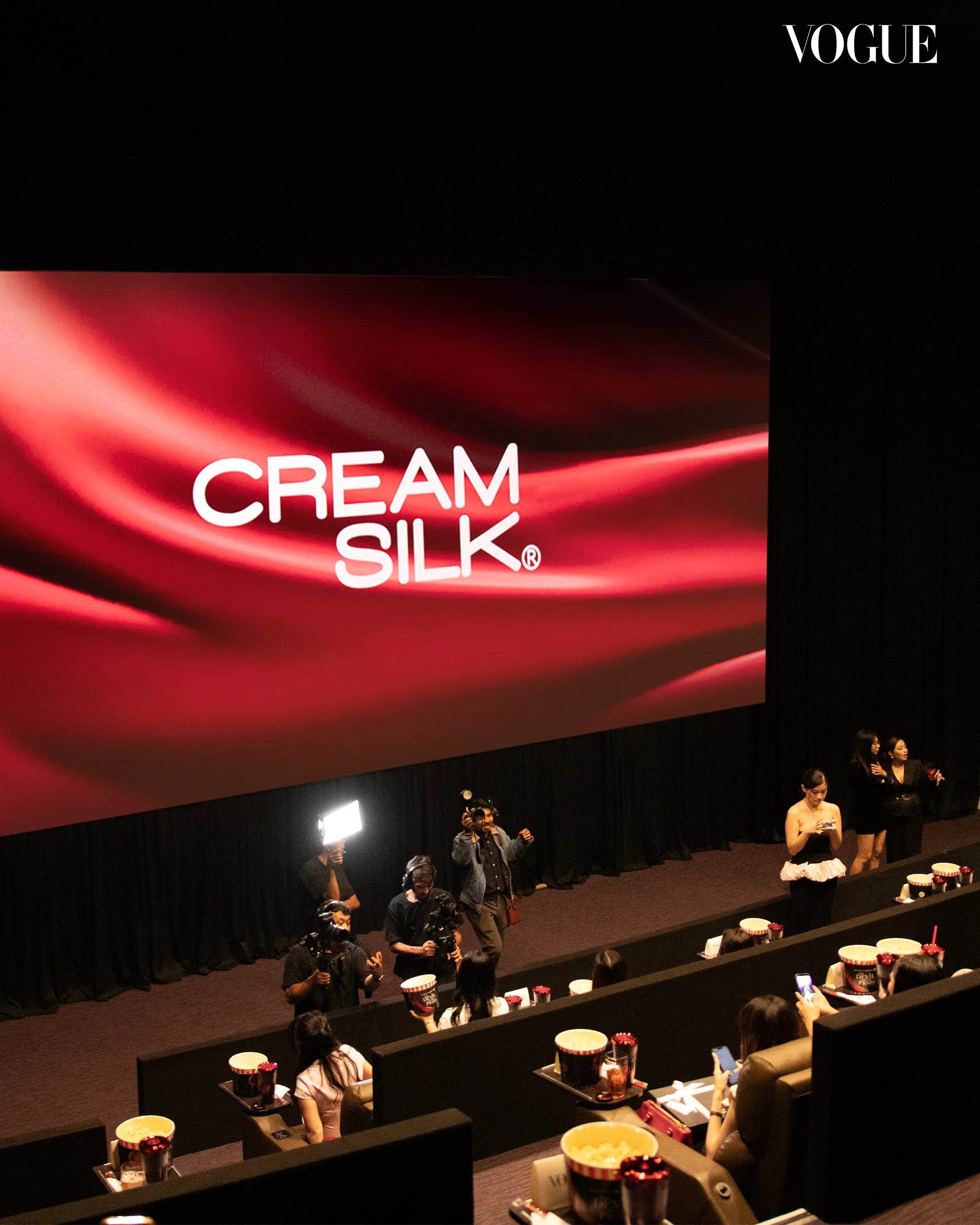 Stage area with a large red backdrop reading 'CREAM SILK' and Vogue logo, photographers and attendees filming and watching from the front rows.