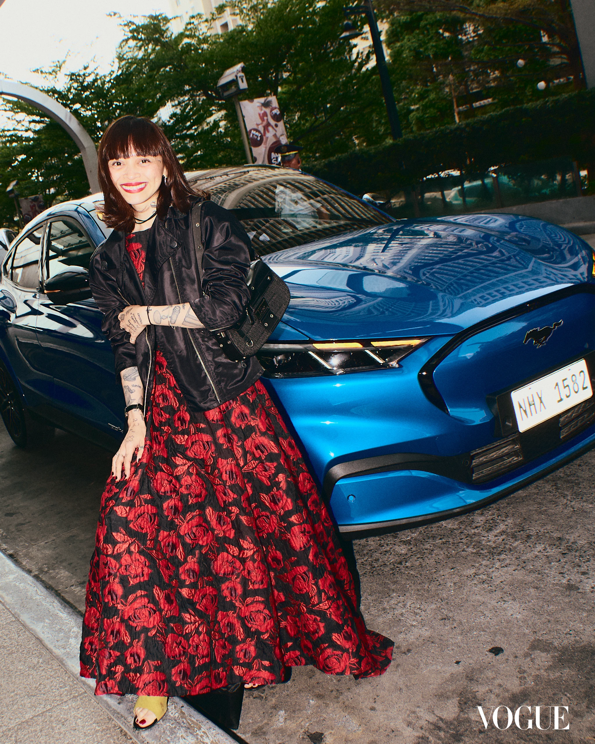 Smiling woman in a red and black floral gown posing beside a blue car on a city street.