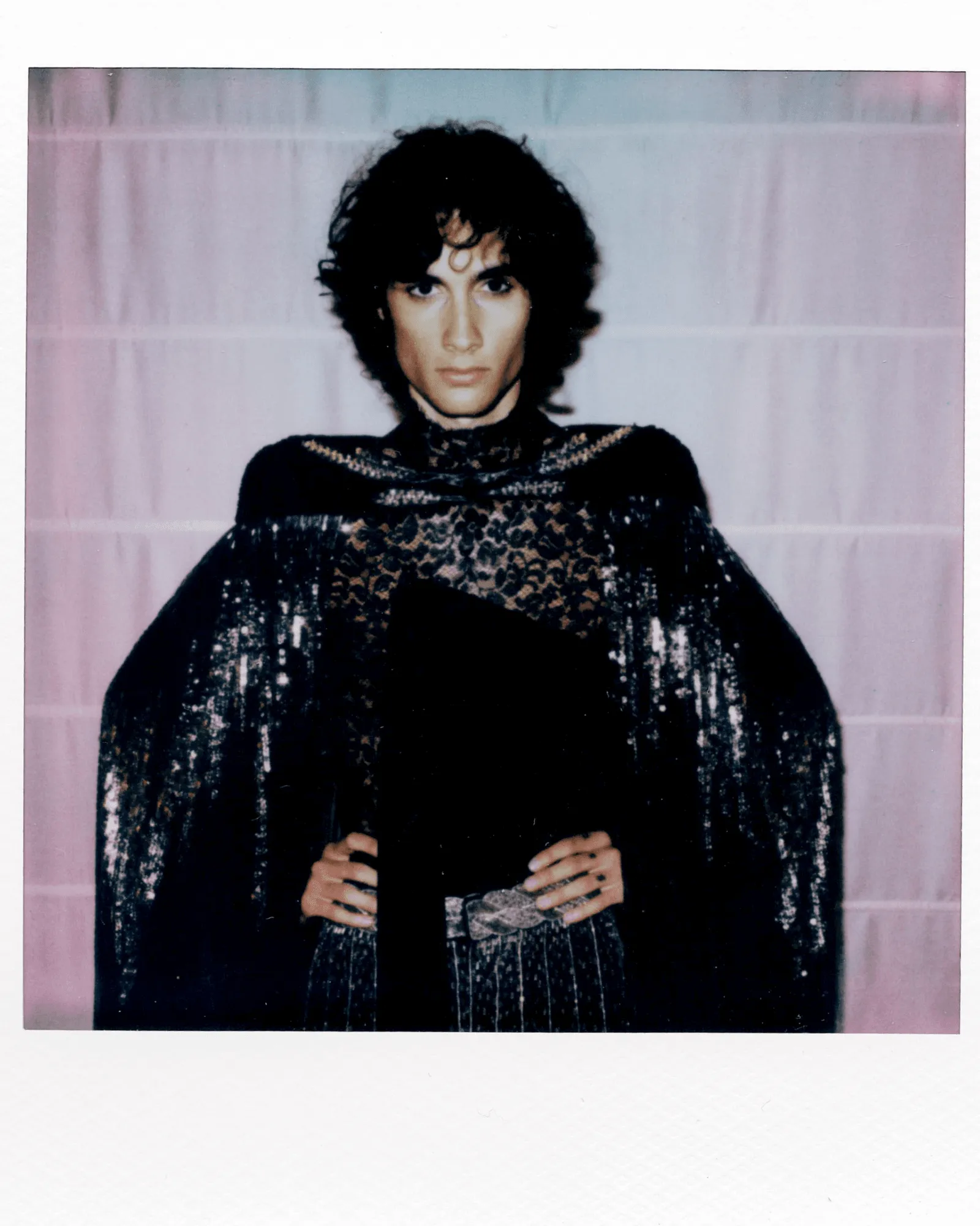Portrait of a person with short dark curly hair wearing a black lace top, standing against a pale pink backdrop, hands on hips.