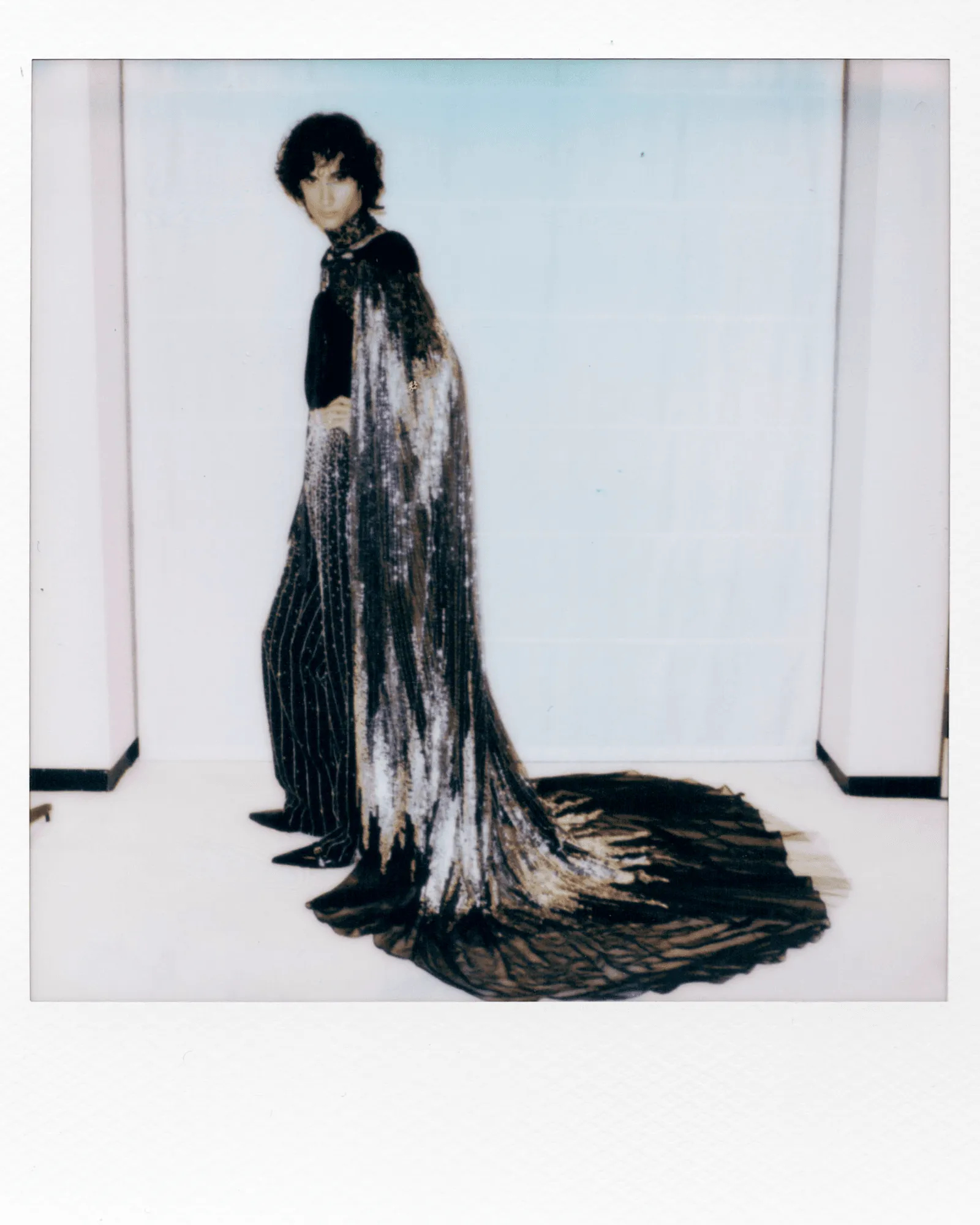 Model wearing a dramatic black and silver floor-length gown with a long trailing train, posed in a white studio backdrop.